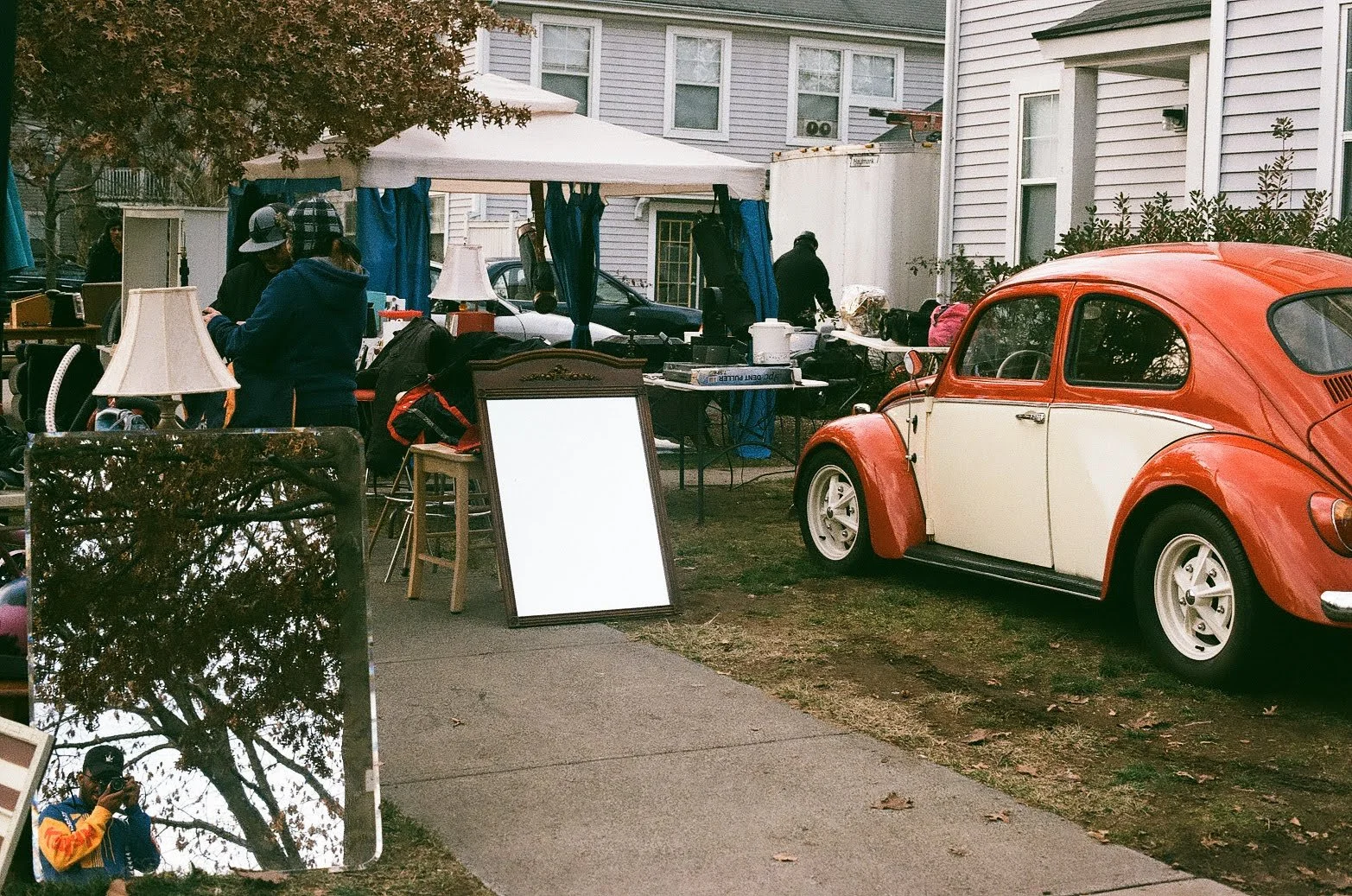 A small vintage orange and white Volkswagen Beetle parked on a grassy area in front of a house with white siding. There is a table with various items, and a large mirror reflecting a person taking a photo of the scene. Several people are browsing ite