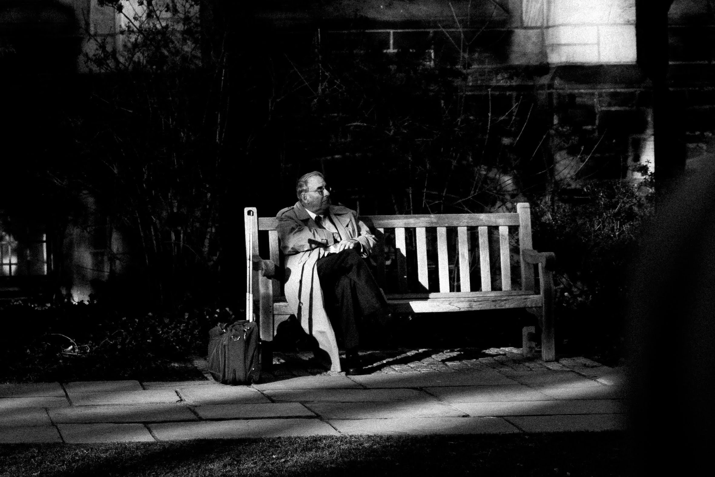 A man wearing glasses and a coat sitting alone on a park bench at night, with a suitcase beside him, illuminated by a nearby streetlight.