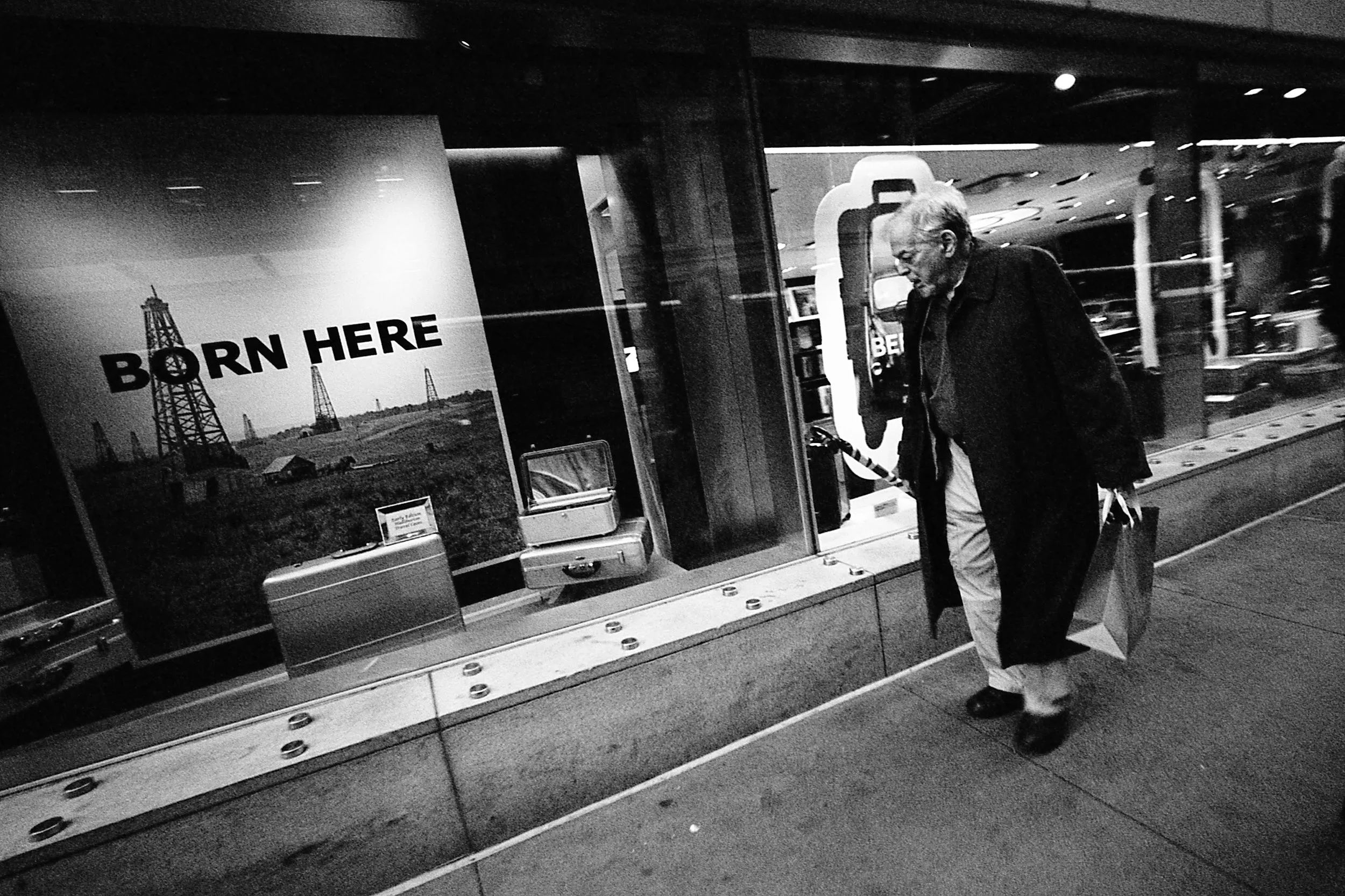 A man carrying a shopping bag and a small suitcase walking in front of an airport window with an advertisement that says 'Born Here' and features oil drills in a rural landscape.