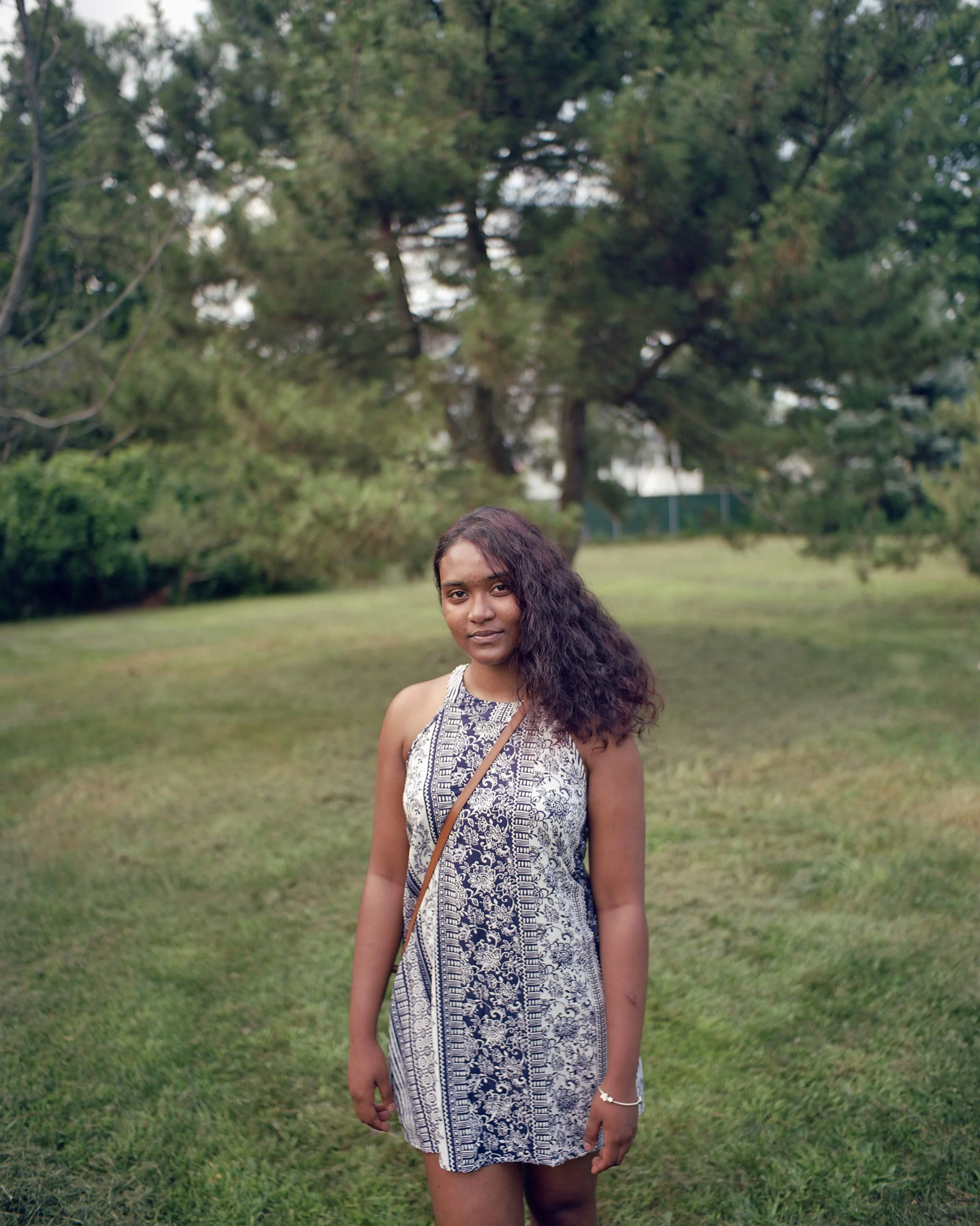 A young woman standing outdoors on a grassy field with trees in the background, wearing a sleeveless patterned dress and carrying a crossbody bag.