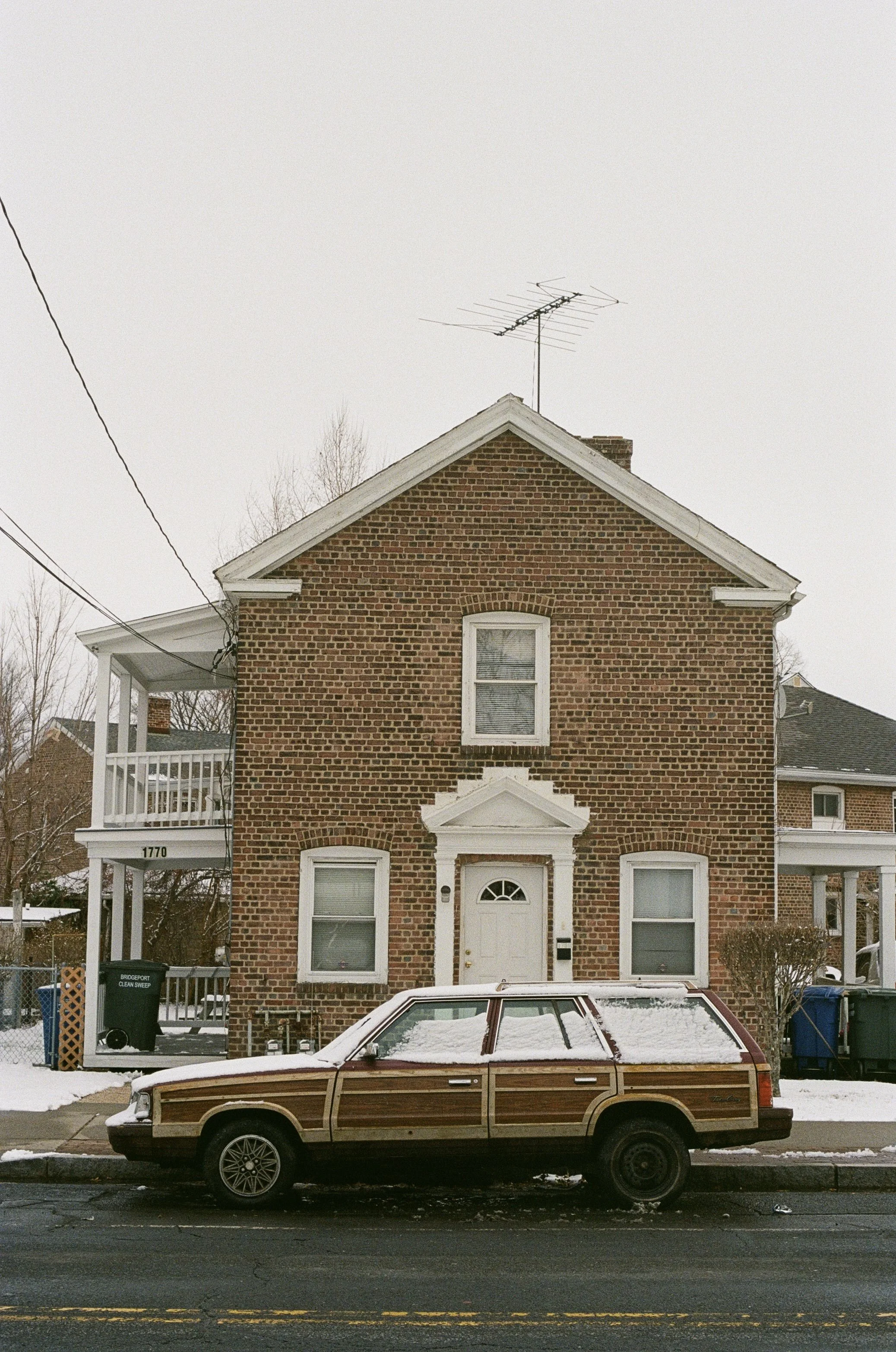 A brick house with two stories and three windows, a white front door, and an antenna on the roof. There's a vintage station wagon with wood paneling parked in front, with some snow on the car. Snow is also on the ground and the sidewalk.