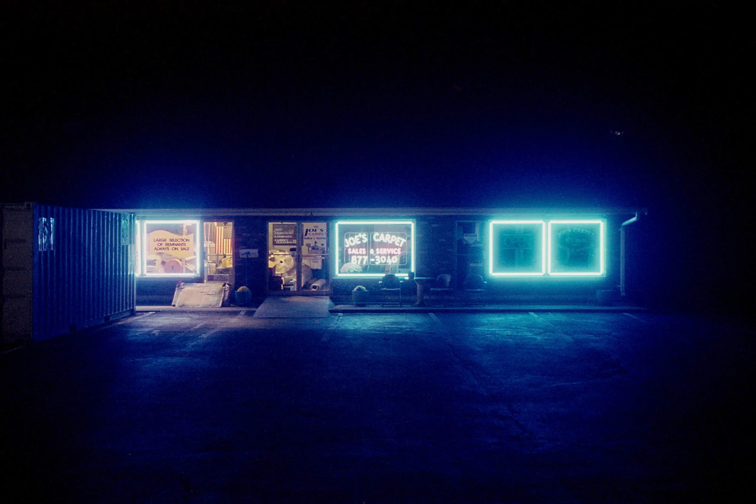 A strip mall storefront at night with neon signs advertising carpet sales and service, with dark sky and minimal surrounding lighting.