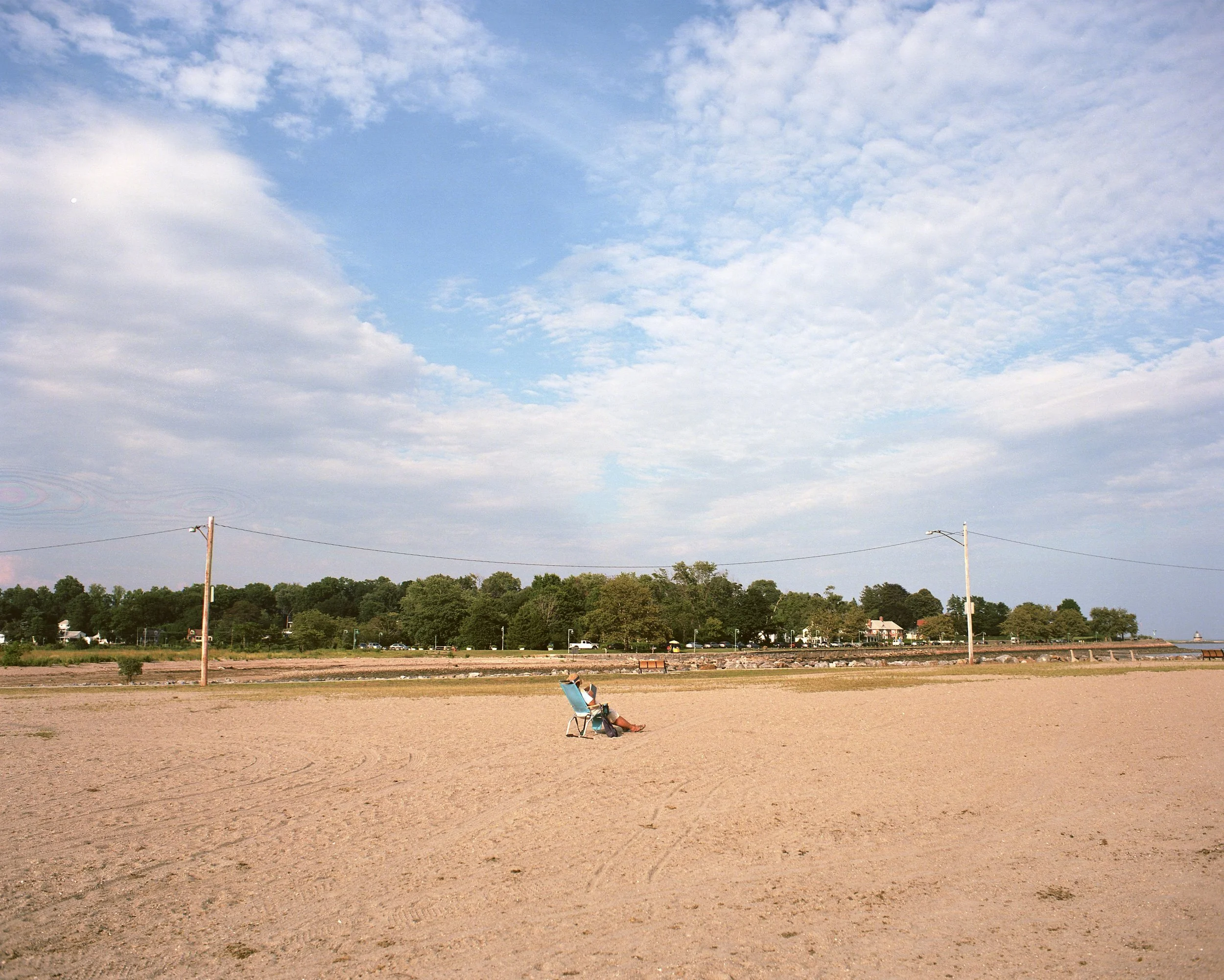 A person sitting on a beach chair on a sandy beach, with a person lying on the sand nearby, under a partly cloudy sky, with trees and houses in the background.