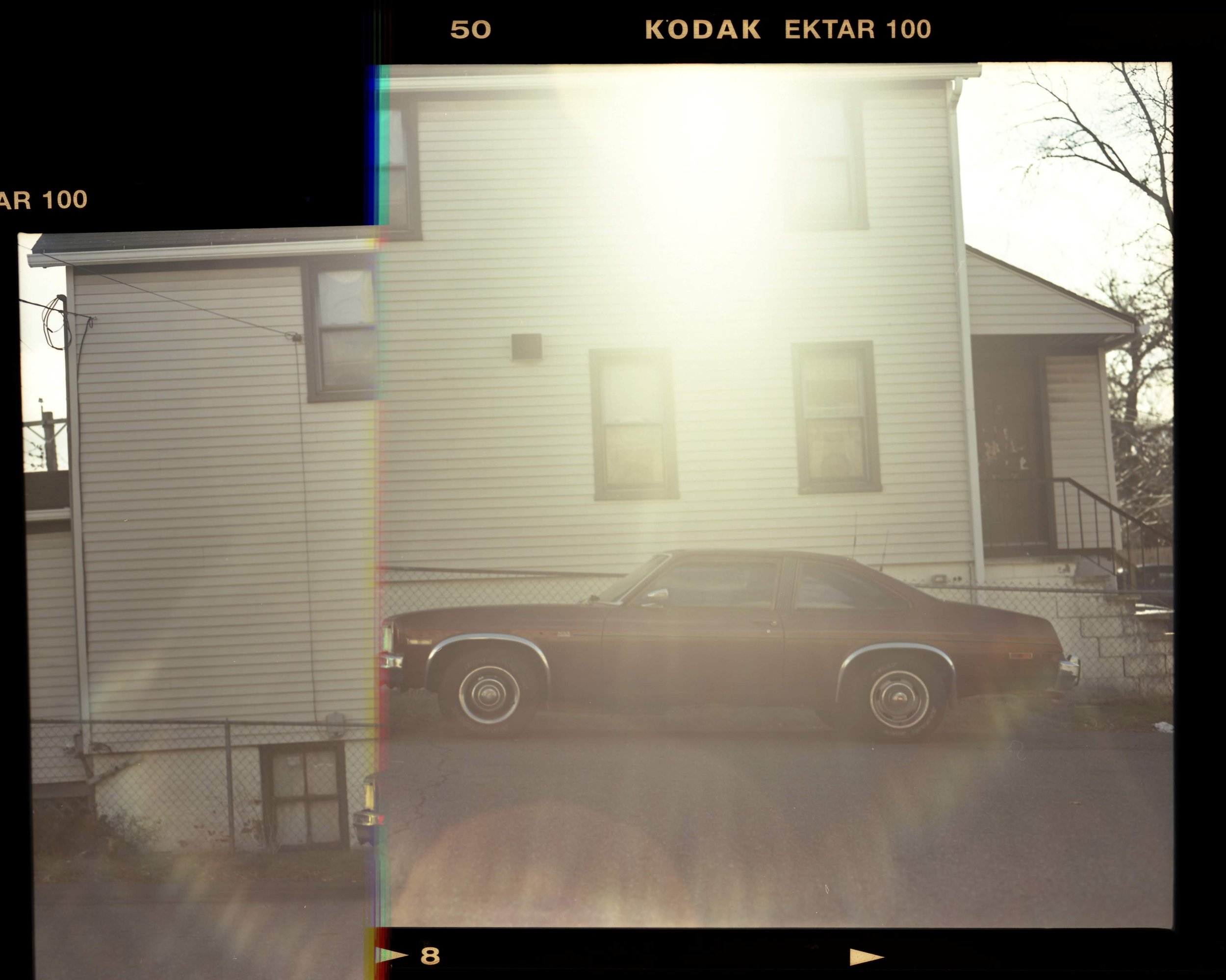 A vintage brown car parked in front of a white residential house with vinyl siding, three visible windows, and a staircase on the right. The photo is overexposed due to bright sunlight.
