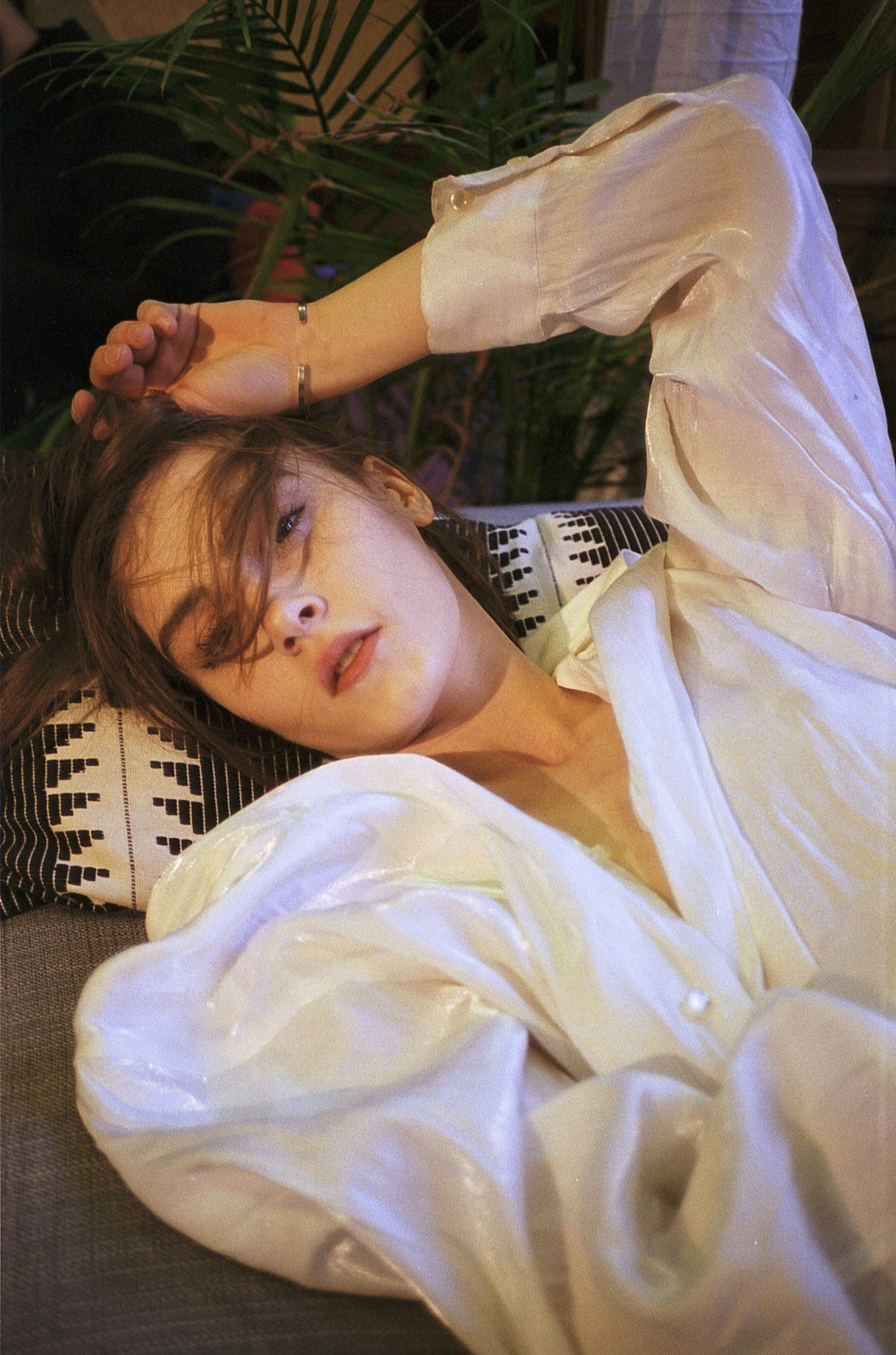 A young woman with medium-length brown hair lying on a patterned pillow, wearing a shiny white blouse, with green plants in the background.