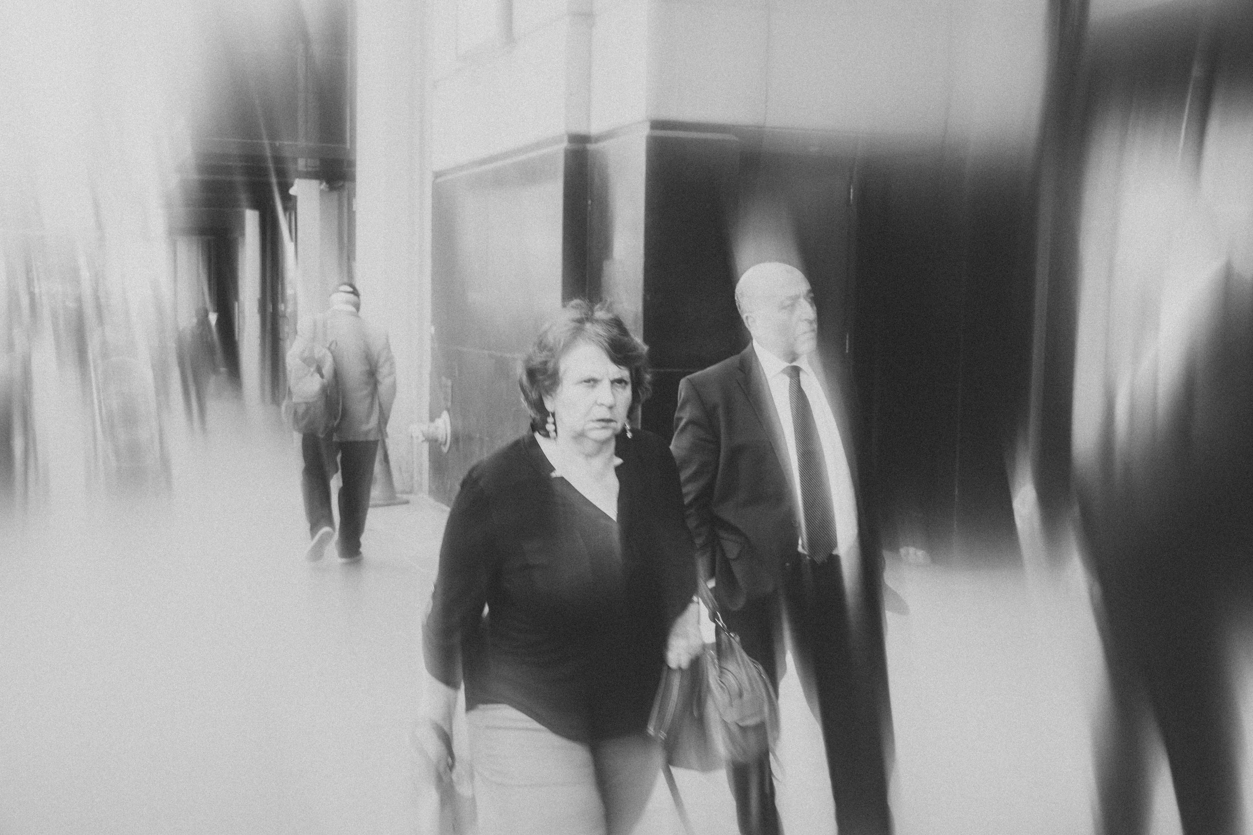 A black and white photo of a woman and a man walking in a hallway with others in the background. The woman has short hair and is wearing a dark top with a purse, while the man is dressed in a suit and tie. The photo appears slightly blurry and has a 