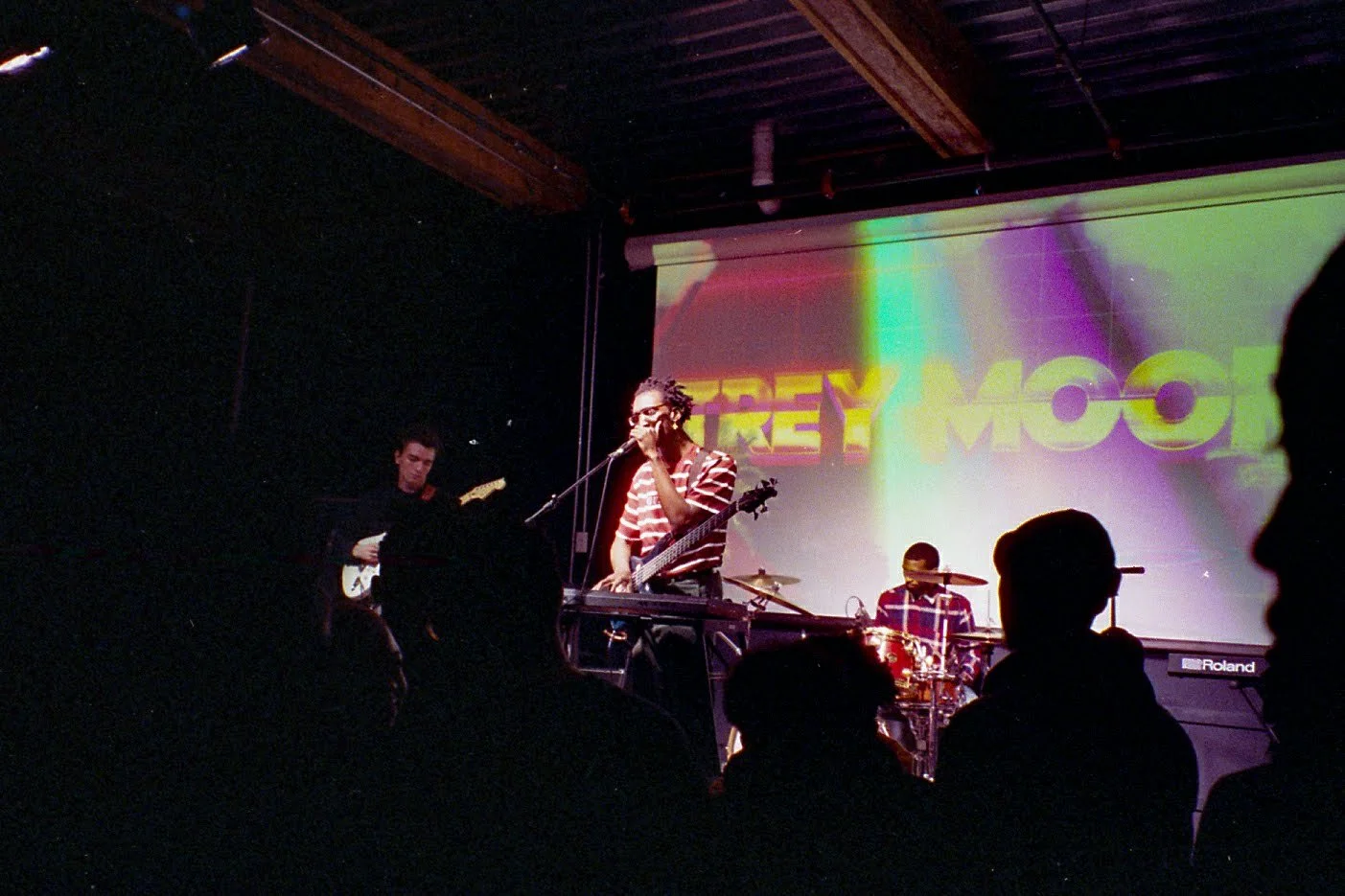 Band performing live on stage with a keyboard, guitar, and drums, in a small venue with an audience watching.