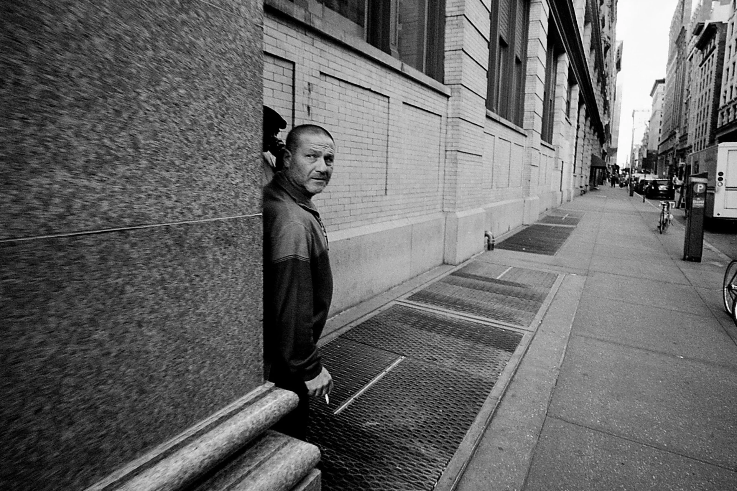 A man with a shaved head and a serious expression, wearing a dark jacket, is standing behind a brick wall on a city sidewalk, looking toward the camera. The scene is in a city with tall buildings, parked cars, bicycles, and pedestrians in the backgro