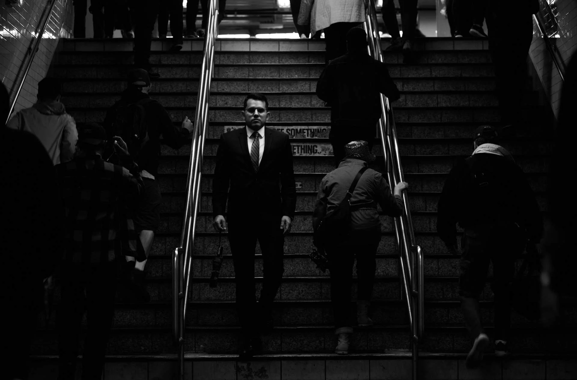 A man in a business suit ascending stairs in a crowded subway station, surrounded by other commuters, with a signed message on the stairs.