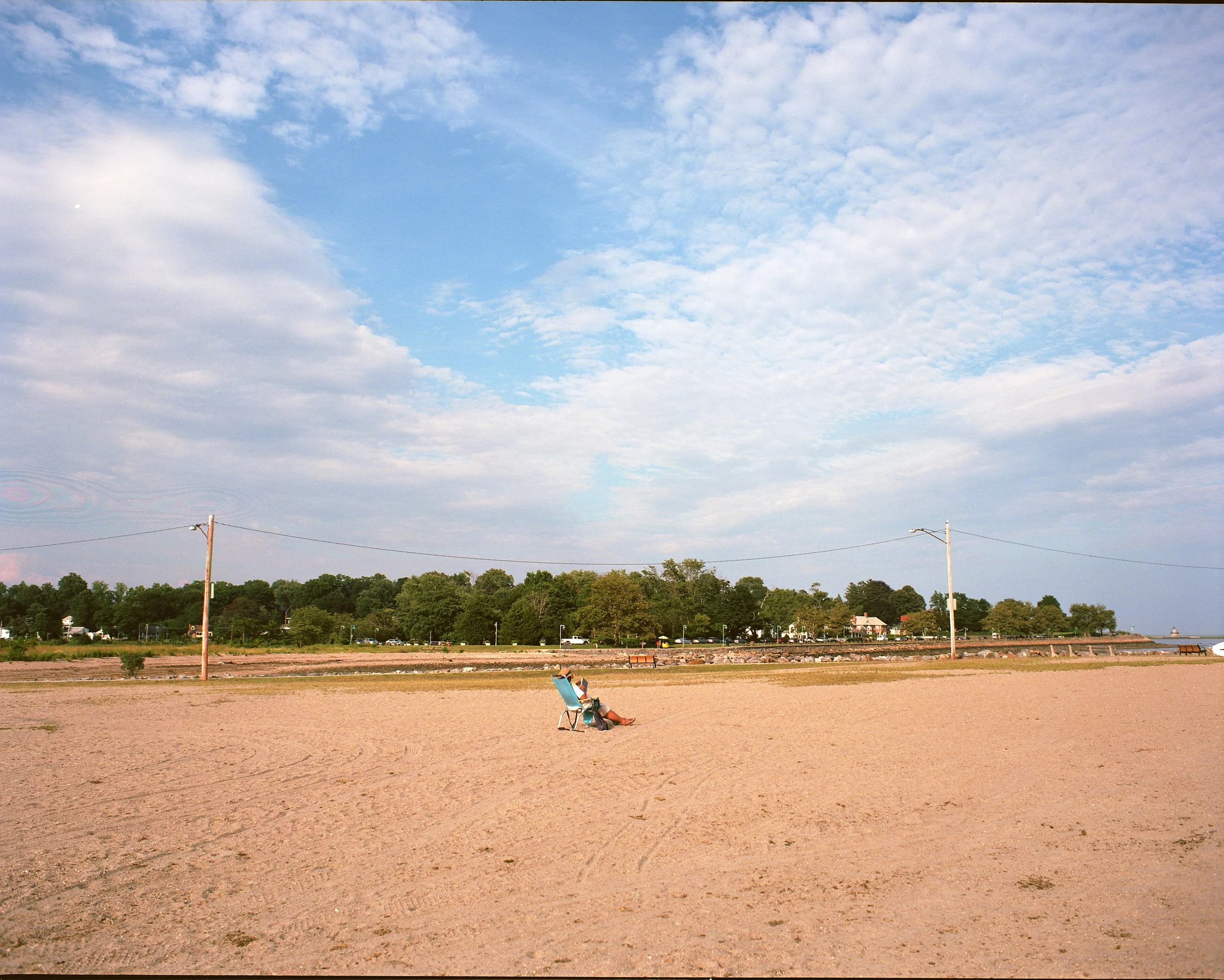 A person sitting on a beach chair on sandy beach under a partly cloudy sky with trees and houses in the background.