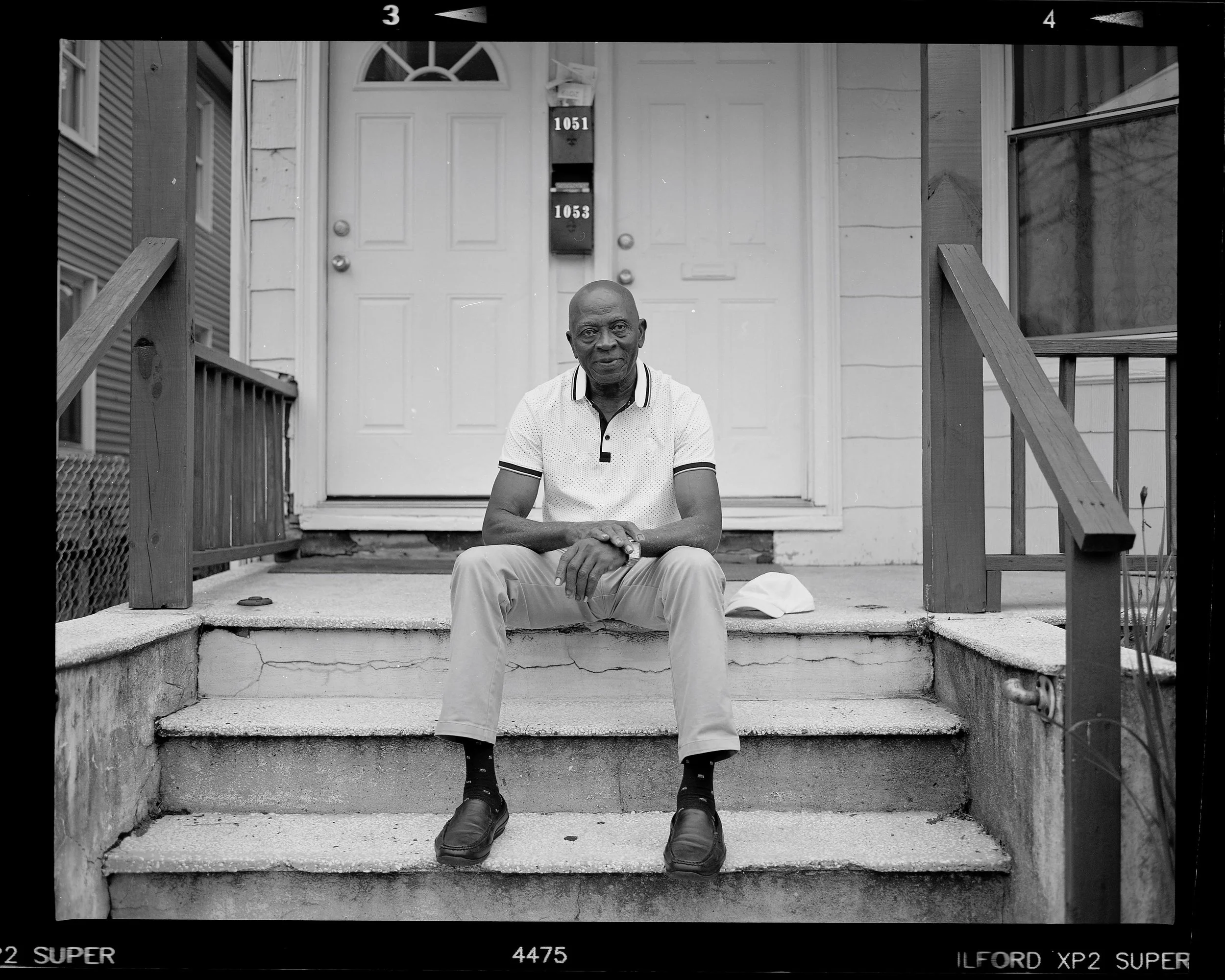 A man sitting on the steps of a porch in front of a white door, with mailboxes and a window nearby, captured in black and white.