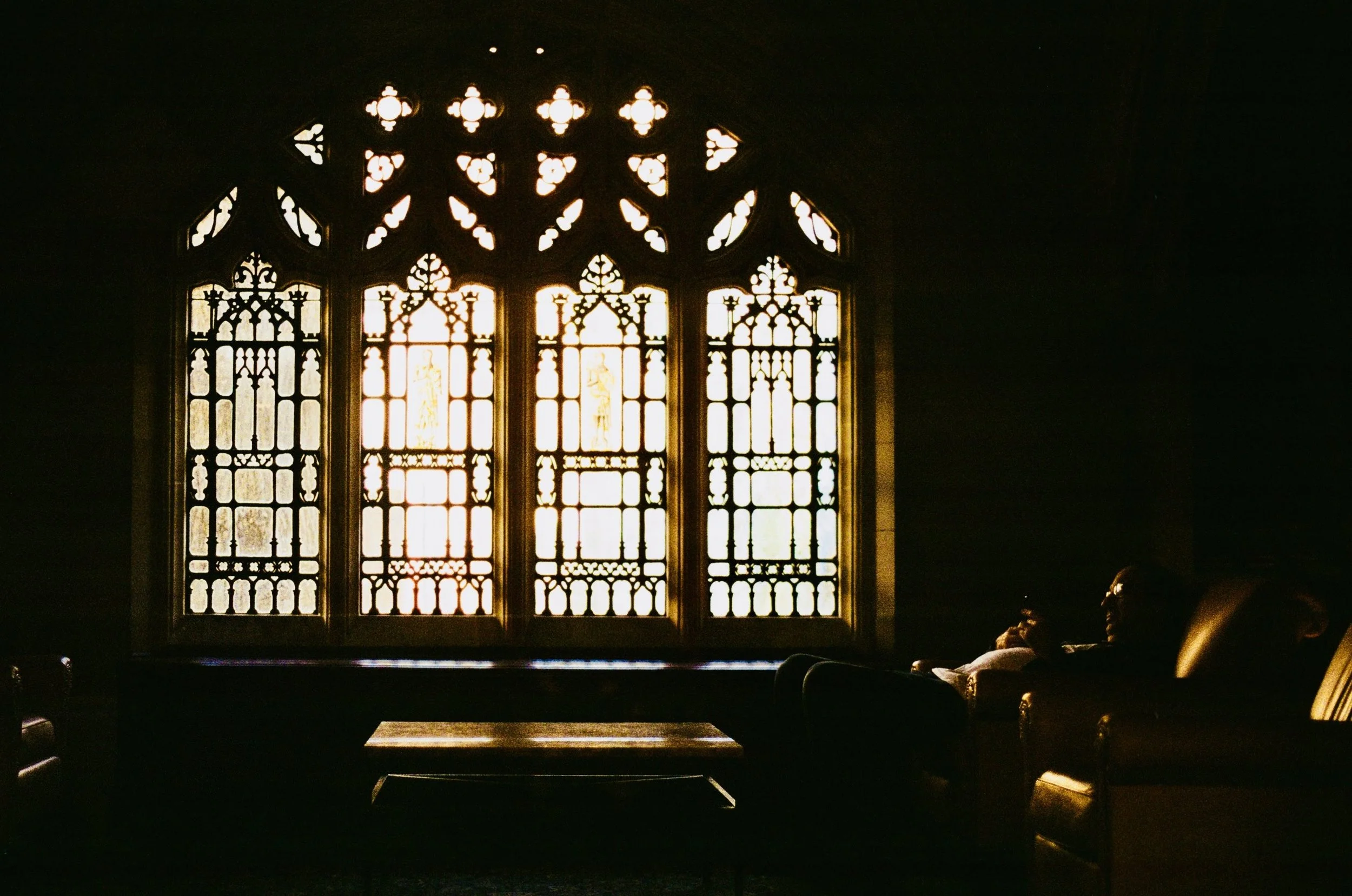 People sitting on chairs and a sofa in a dark room, with large Gothic-style stained glass windows allowing sunlight to stream in.