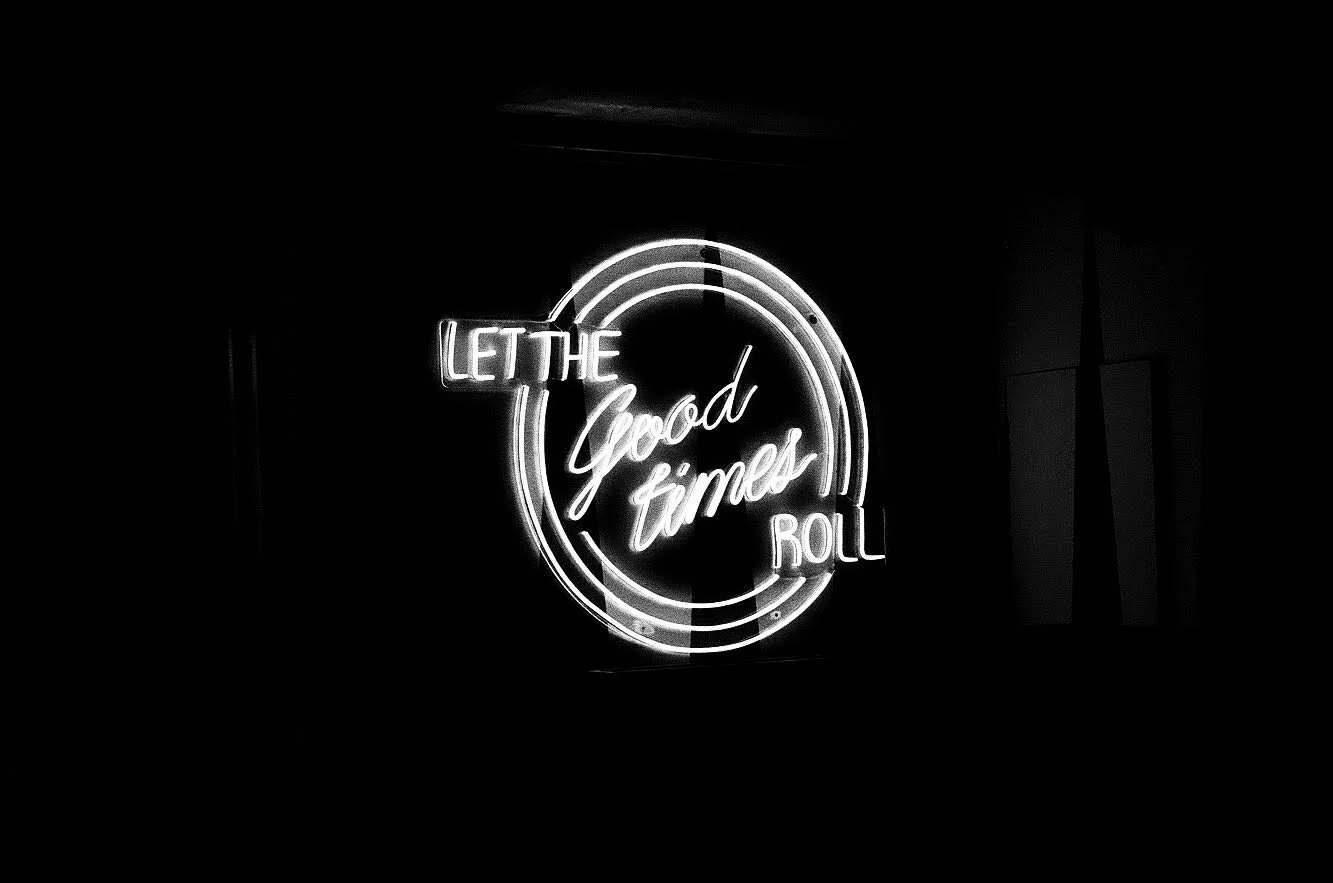 Neon sign that says "Let the good times roll" with a circular design around the text, glowing in white on a dark background.