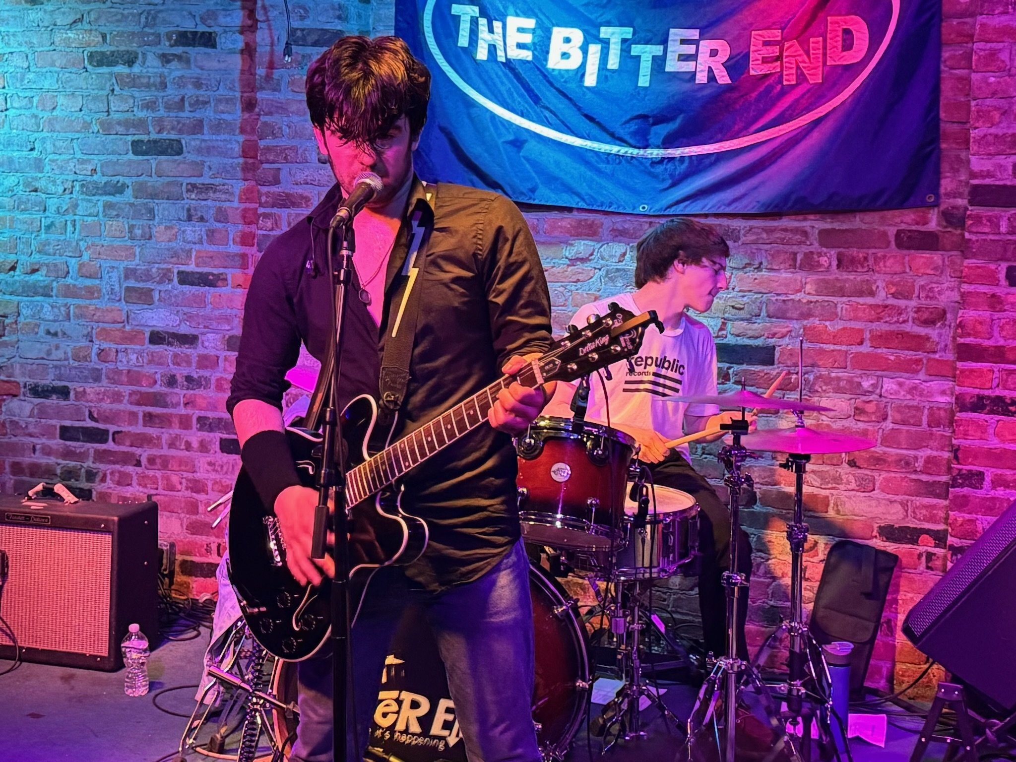 Two young male musicians perform on stage with a brick wall background. The one in the foreground plays an electric guitar and sings into a microphone, wearing a black shirt and jeans. The one in the background plays drums, wearing a white t-shirt with text, and glasses. A blue banner with the words "THE BITTER END" hangs behind them.