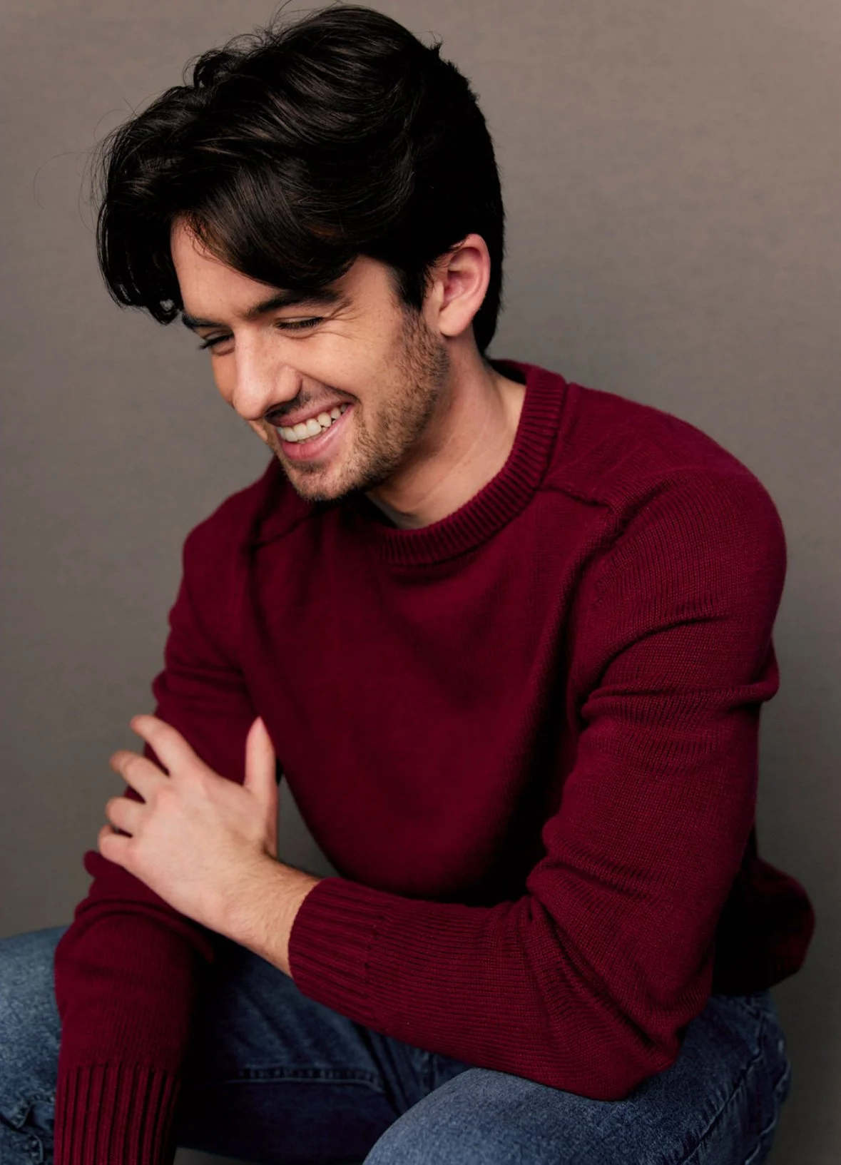 A young man with dark hair and a beard, sitting against a gray background, wearing a red sweater, smiling and looking down with his eyes closed.