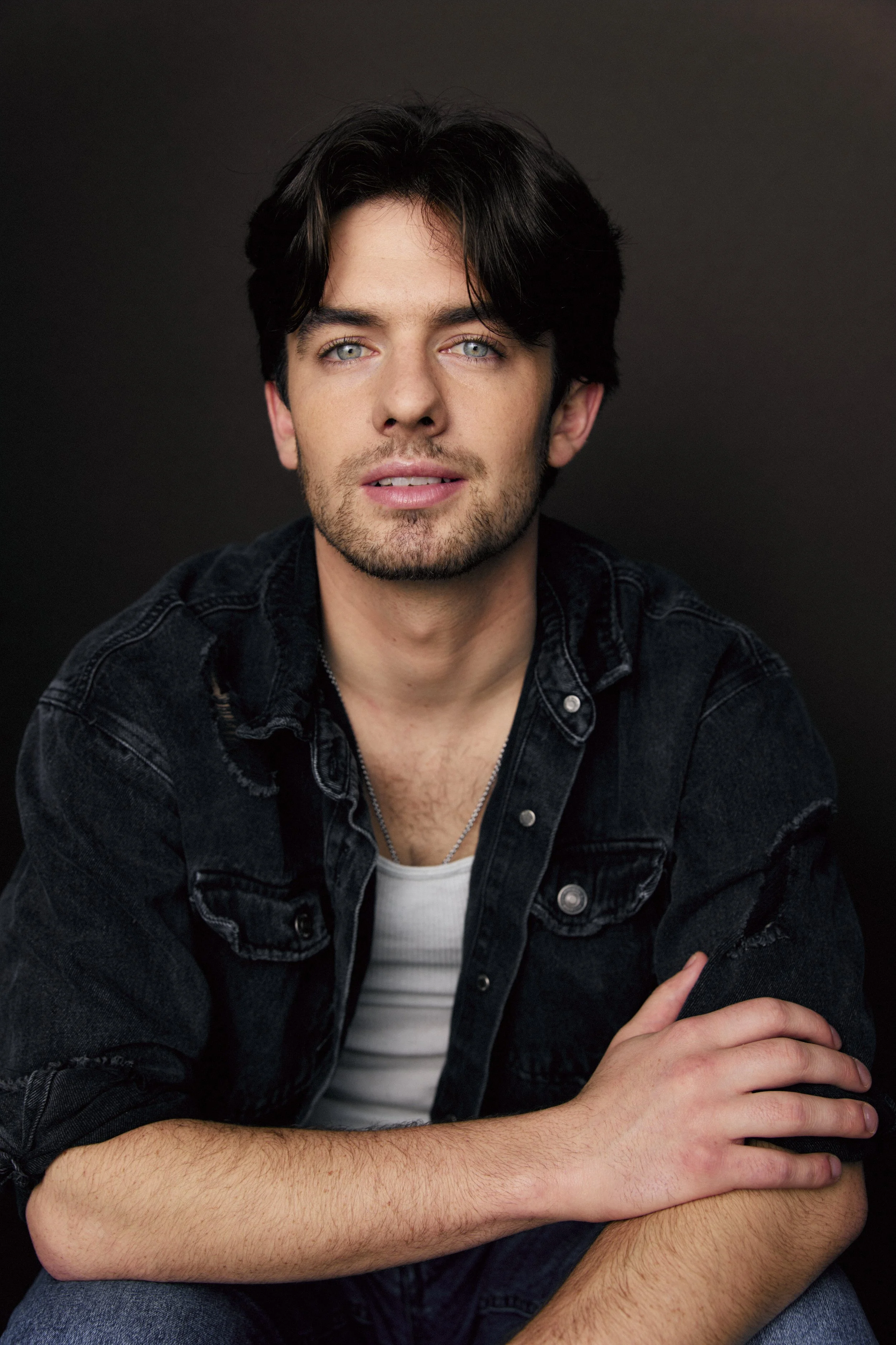 A young man with dark hair, blue eyes, and light skin, wearing a black denim jacket and a white shirt, seated against a dark background.