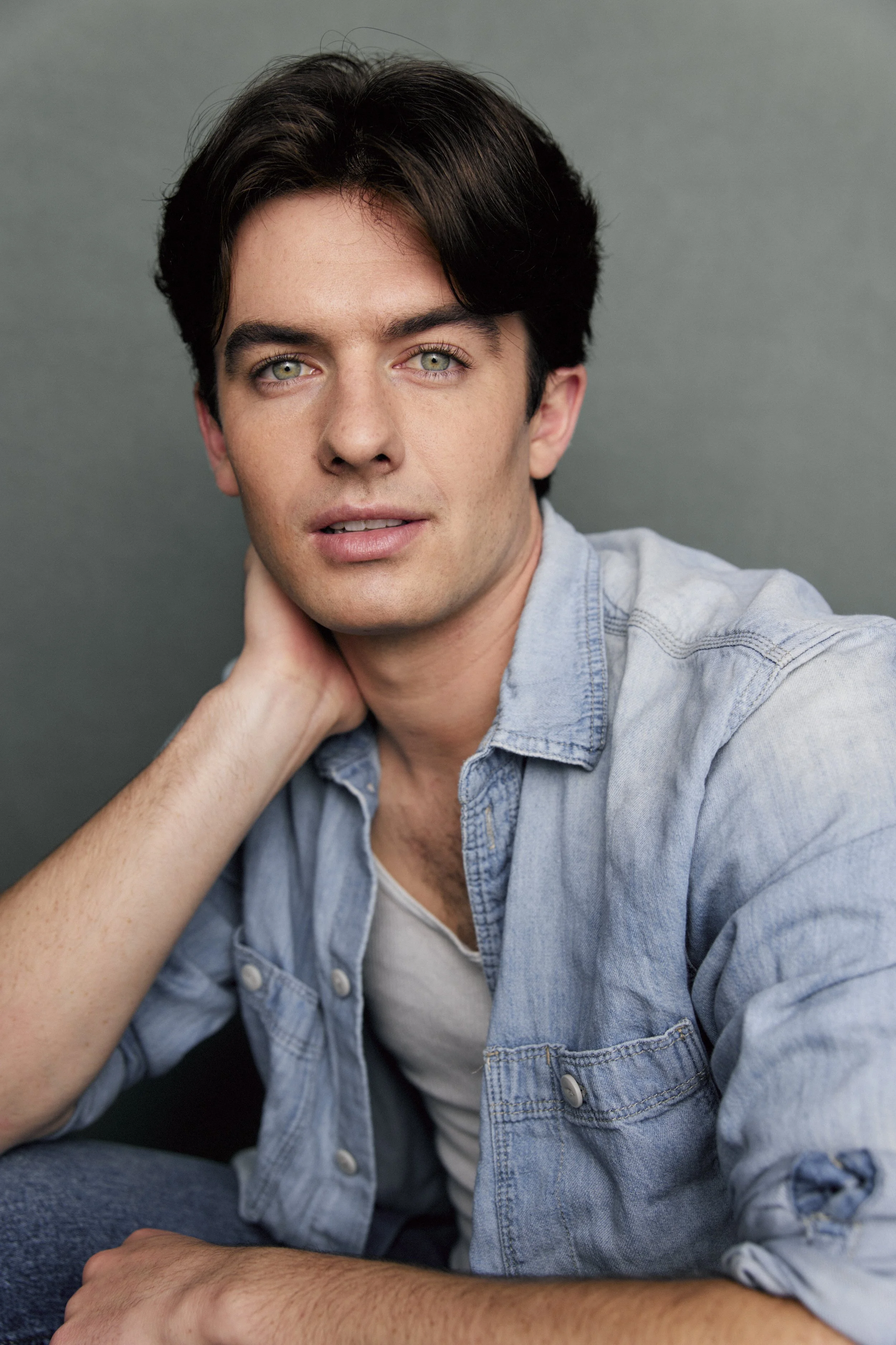 A young man with dark hair and light-colored eyes, wearing a denim shirt and white t-shirt, sitting against a gray background and looking at the camera.