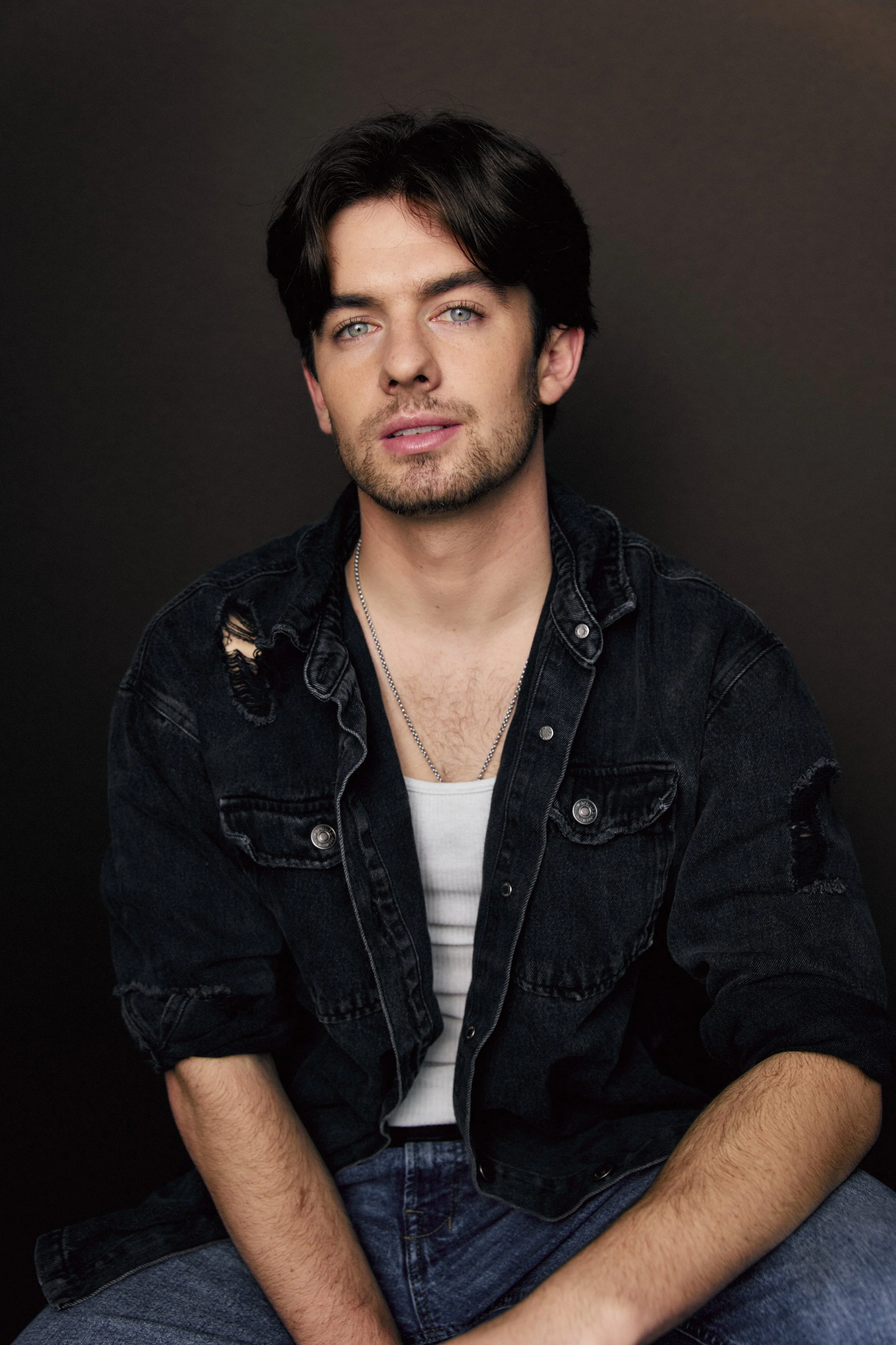 A young man with dark hair, blue eyes, and light skin, wearing a ripped black denim jacket over a white shirt, sitting against a dark background.