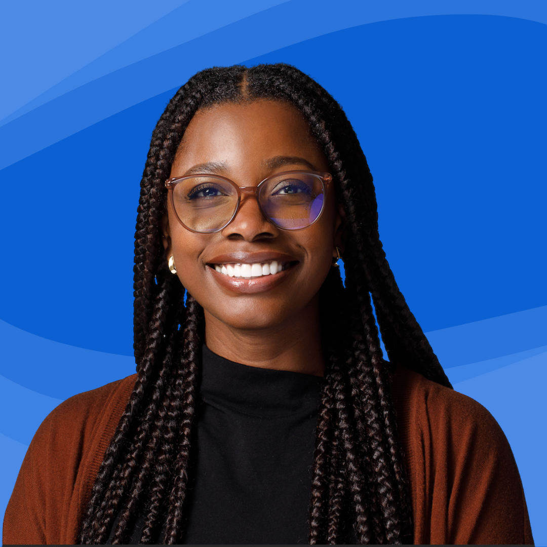 Portrait of a smiling Nigerian woman with long braided hair wearing glasses, earrings, a black shirt, and a brown jacket against a blue background.