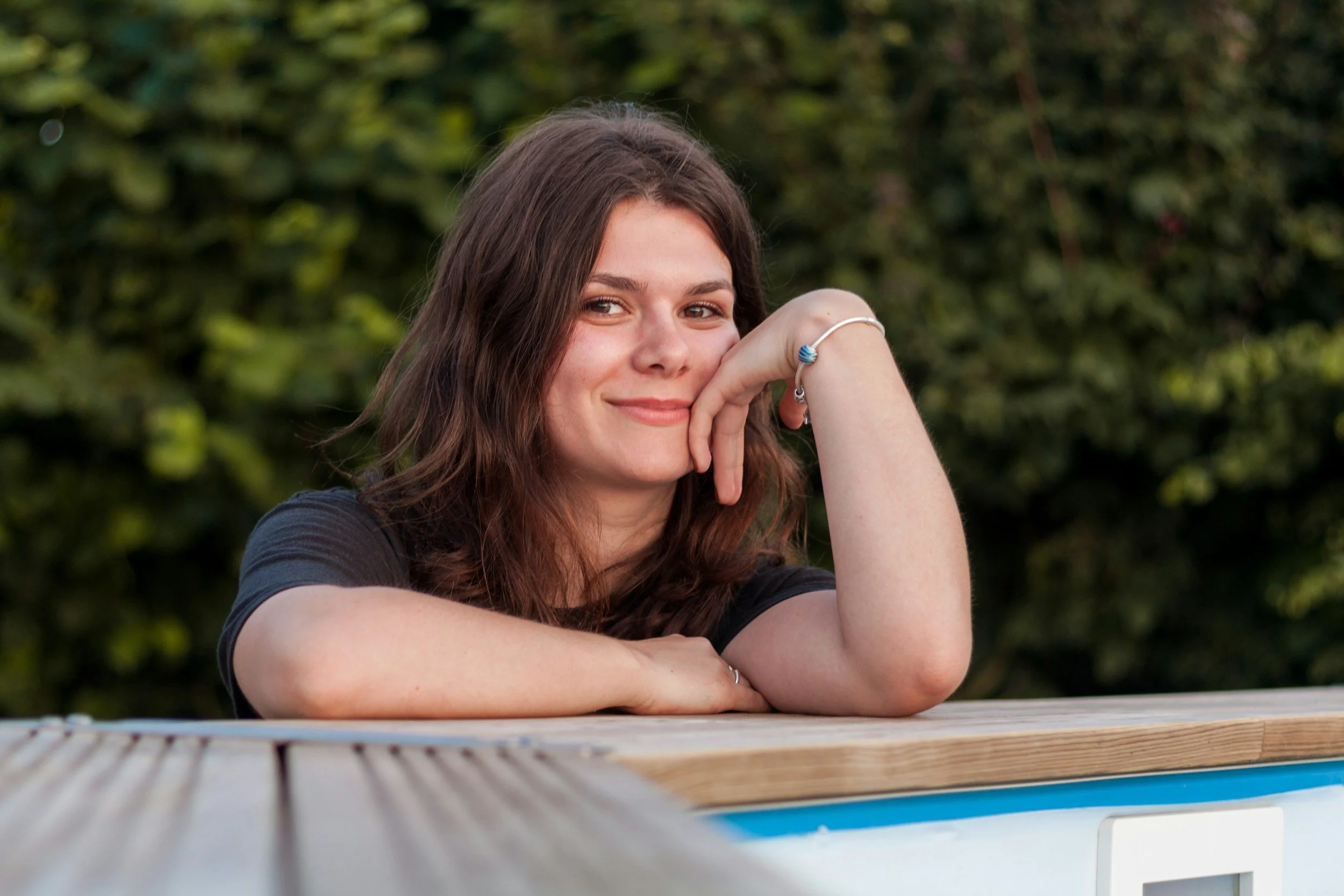 A young woman with brown hair smiling, resting her chin on her hand, sitting at a wooden table outdoors with green foliage in the background.