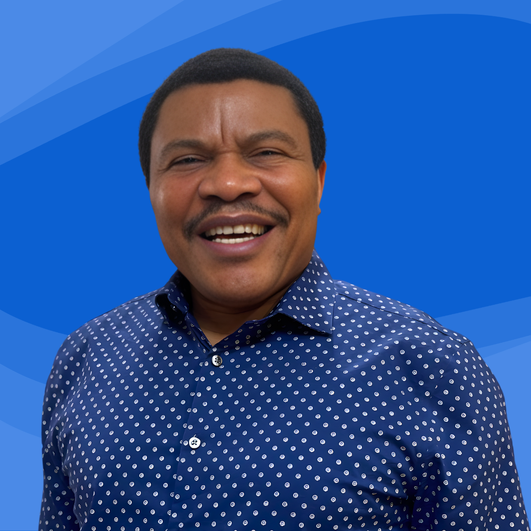 A man with dark hair, wearing a blue dress shirt with white circular patterns, smiling with his teeth showing, against a blue background with lighter blue curved lines.