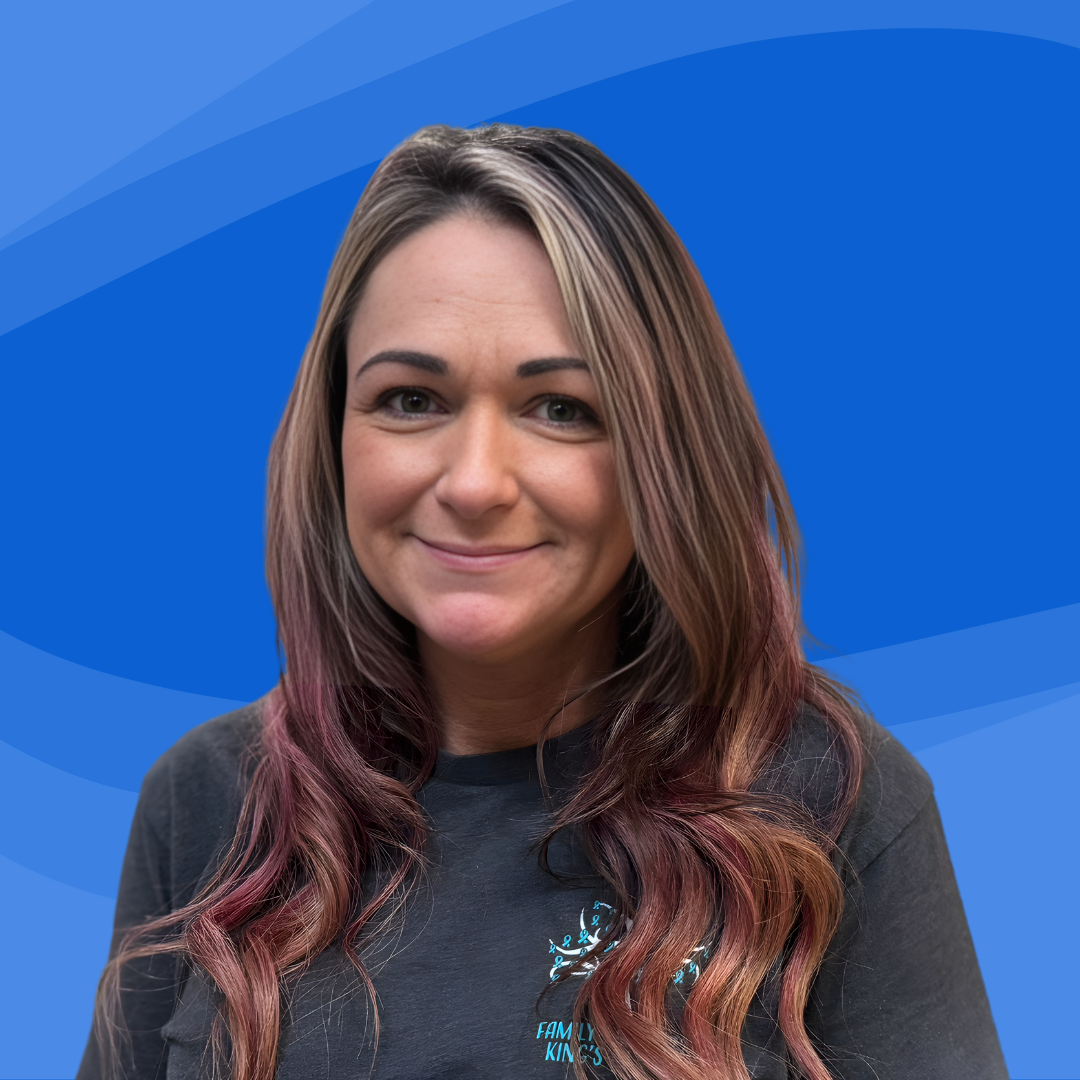 A smiling woman with long, wavy hair dyed with pink and purple highlights, wearing a black T-shirt with a logo that says 'Family Kindness' on it, against a blue background.