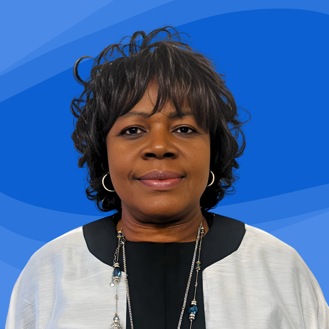The clinic director, a Nigerian woman with short dark hair wearing hoop earrings, a black and white blouse, and layered necklaces against a blue abstract background.