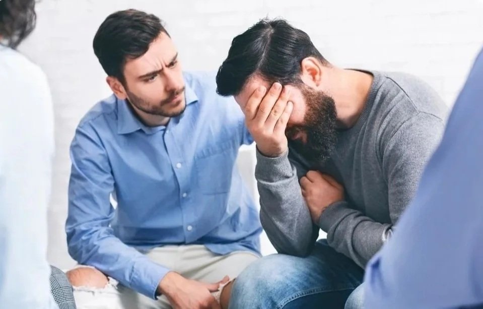 A man with a beard is sitting on a chair, holding his face in his hand, appearing distressed. Another man is sitting close by, gently touching his shoulder or arm, showing concern. They seem to be having an emotional or serious conversation.