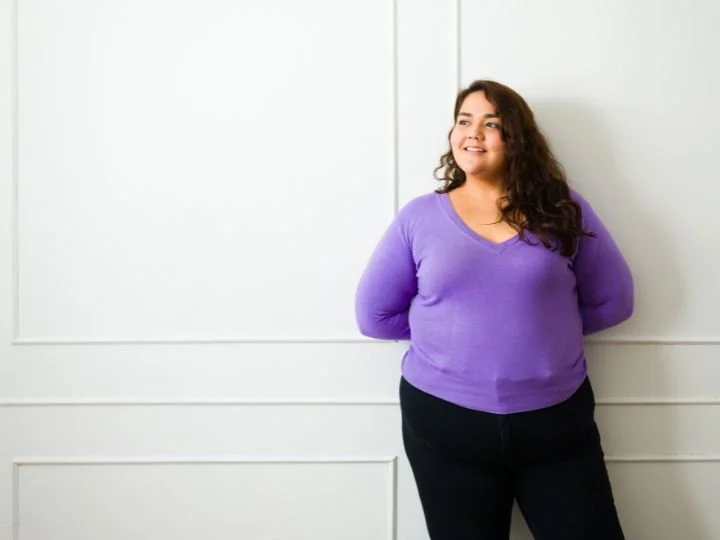 Woman with dark curly hair wearing a purple sweater and black pants standing against a white wall, smiling.
