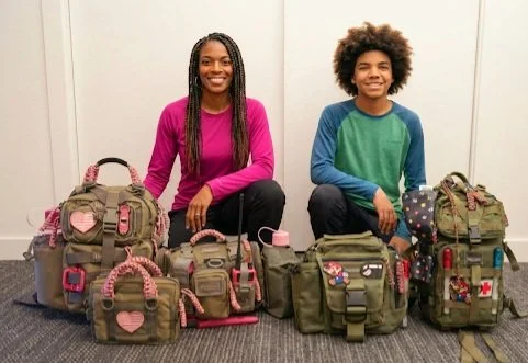Two people, a woman and a young boy, sitting on the floor with bags and backpacks decorated with patches and accessories.