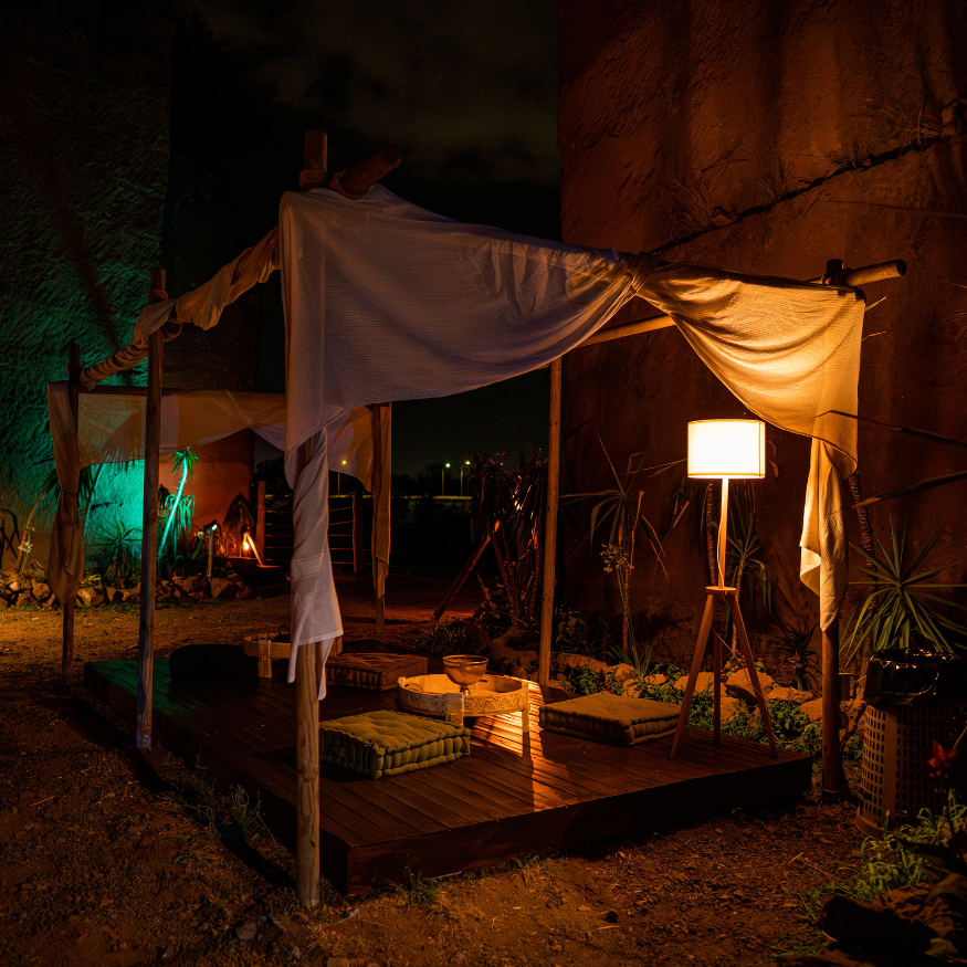 Zona relax all'aperto con tavolinetto, cuscini e lampada, illuminata da luci calorose e luci colorate, di notte.