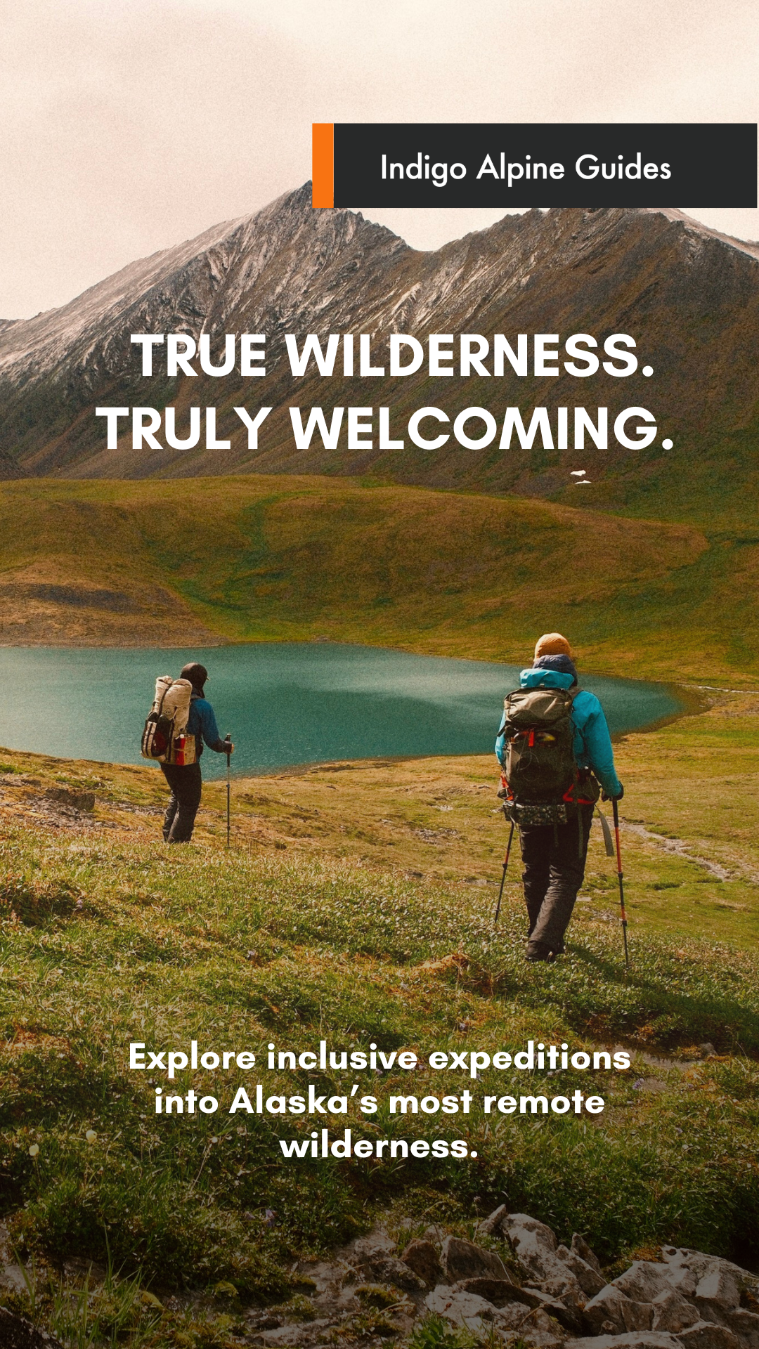 Two hikers trek through a grassy landscape near a mountain and lake in Alaska.