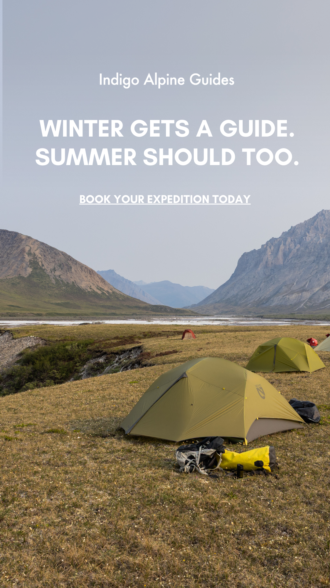 Tents set up in a grassy valley surrounded by mountains, with a river running through the background.