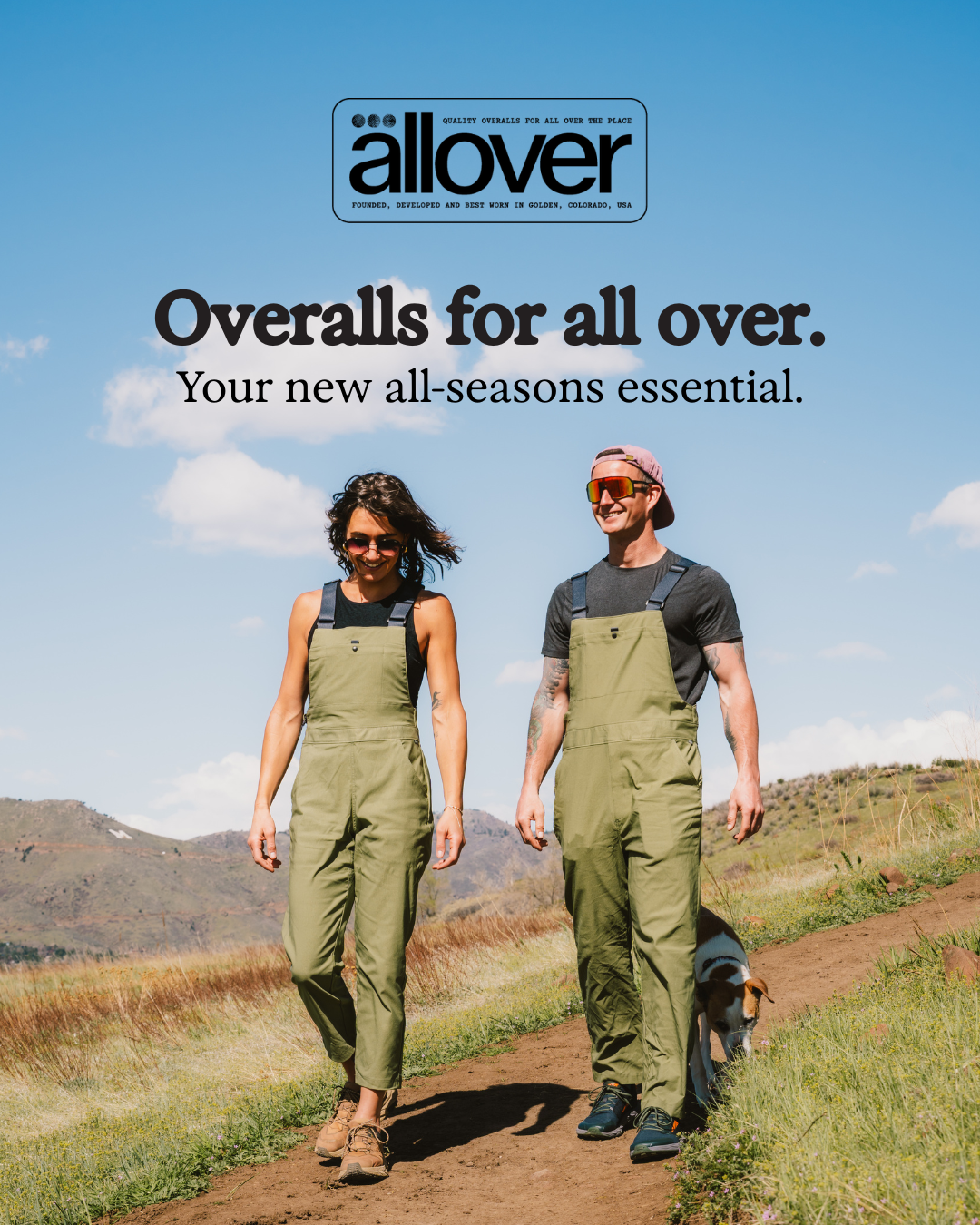 Two people wearing green overalls walking outdoors on a dirt trail with mountainous landscape, a woman with curly hair and a man with tattoos and a pink cap, smiling, accompanied by a dog, under a clear blue sky with some clouds.