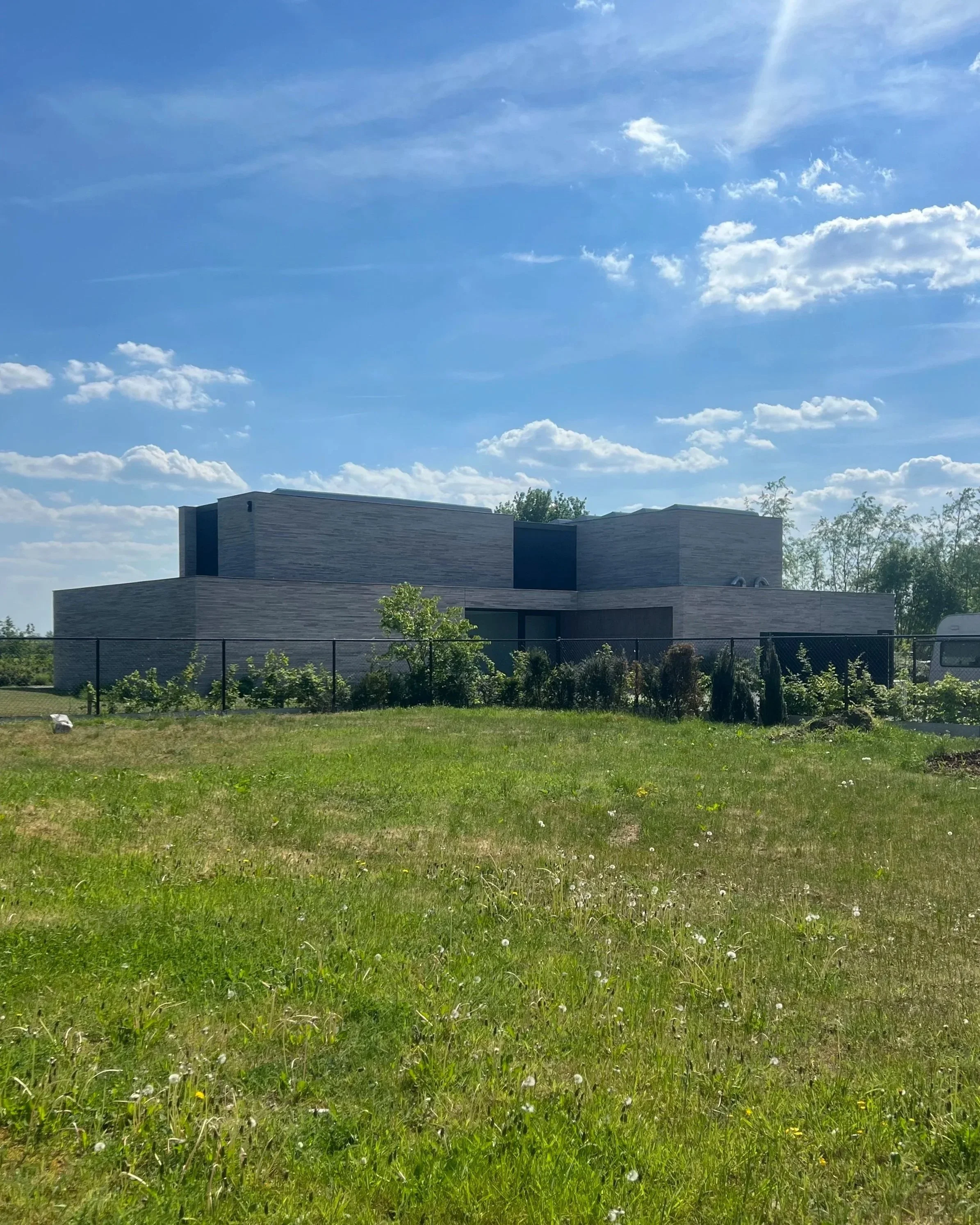 Modern building with flat roof and horizontal siding, partially obscured by a grassy lawn with small plants and a black fence in the foreground, under a bright blue sky with scattered clouds.