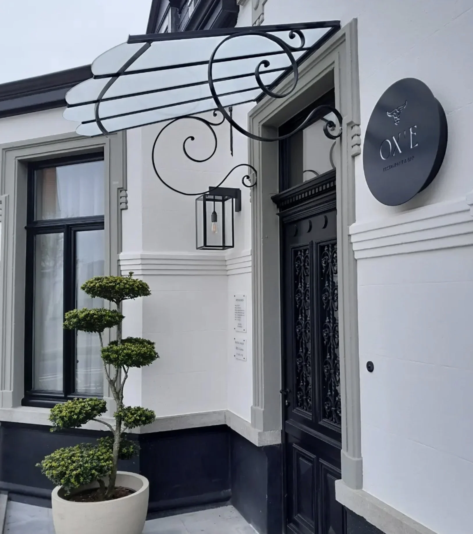 Modern building entrance with black ornate door, white walls, and a sign reading 'OXE restaurant & bar'. A decorative black metal canopy and lantern light fixture are above the door, and a potted topiary tree is in front.