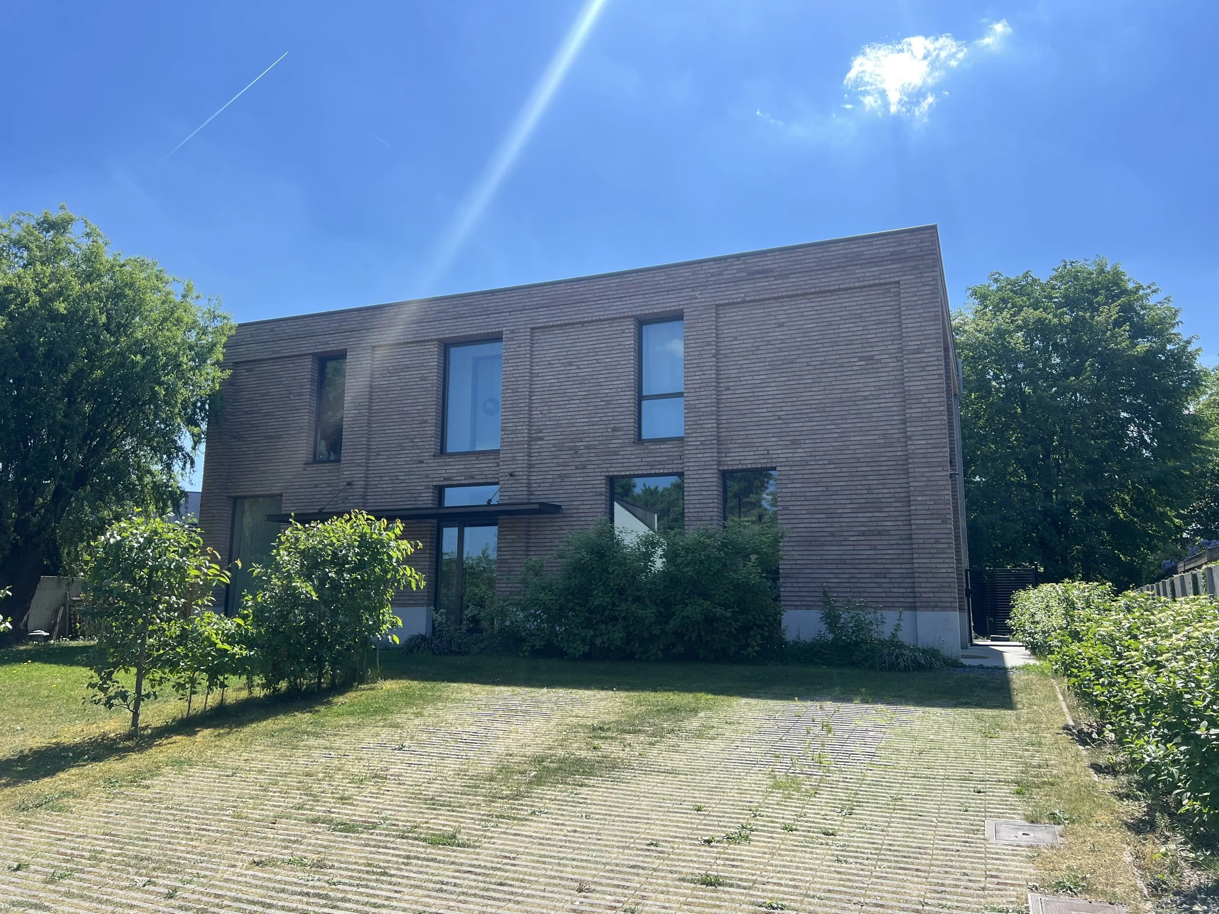 A modern brick building with large windows, surrounded by green trees and bushes, under a bright blue sky with a sunbeam and a contrail.