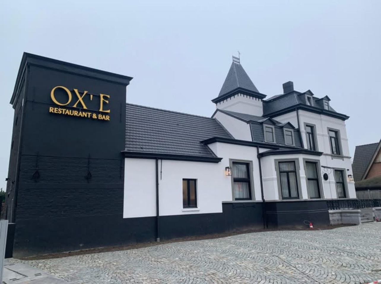 A building with a sign that reads "Oxe Restaurant & Bar," combining a traditional mansion style with black and white exterior, featuring a turret, multiple windows, and a cobblestone driveway.