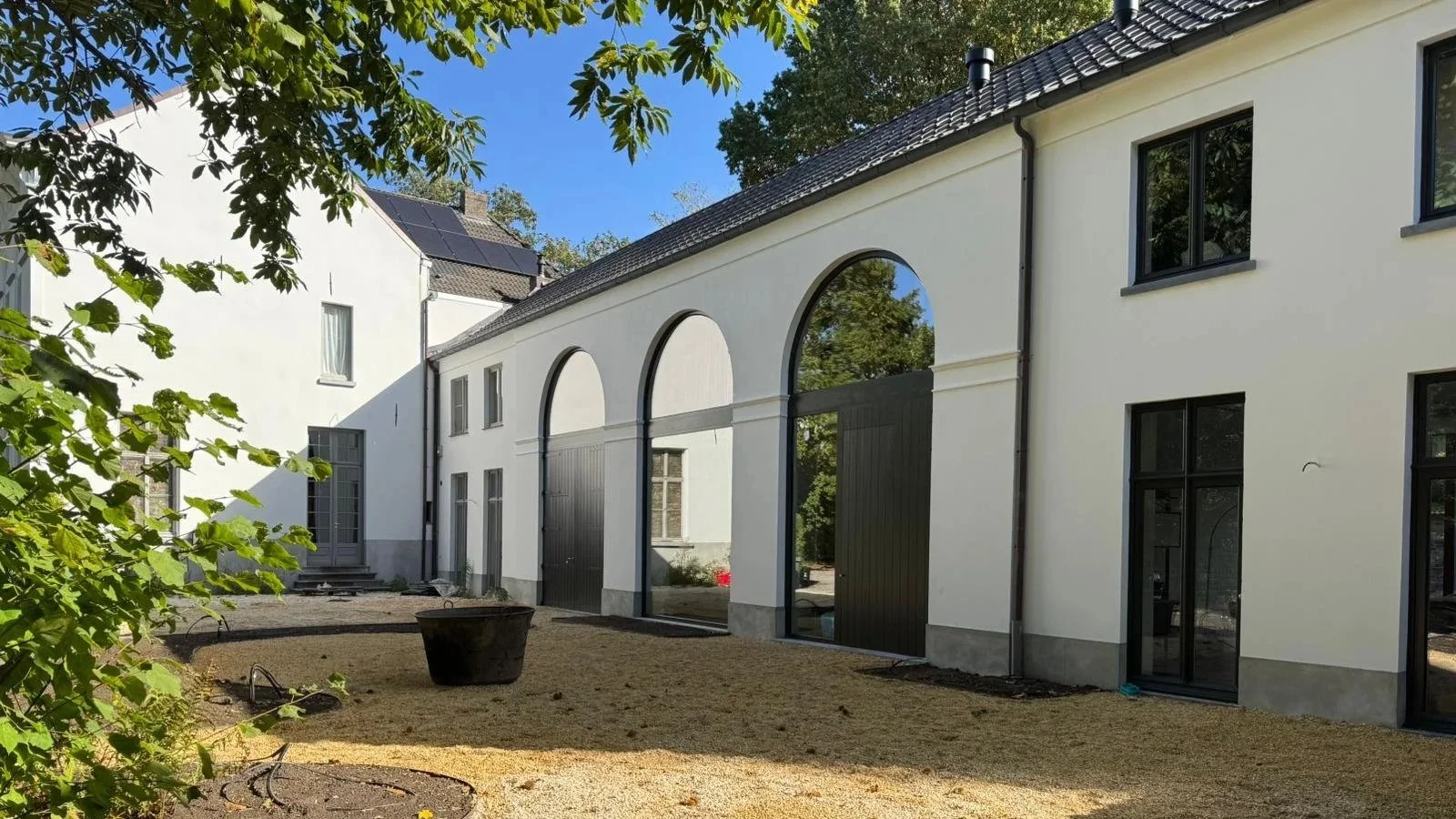 Modern white building with black window frames and arched mirrors in the doors, outdoor area with gravel and trees, clear blue sky.