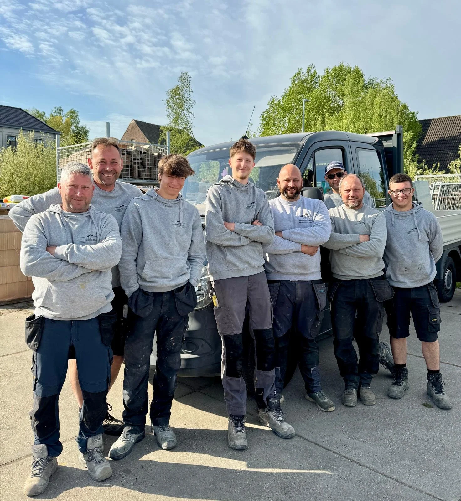 Group of eight men in work clothes standing in front of a work truck, smiling with arms crossed, outdoors on a sunny day.
