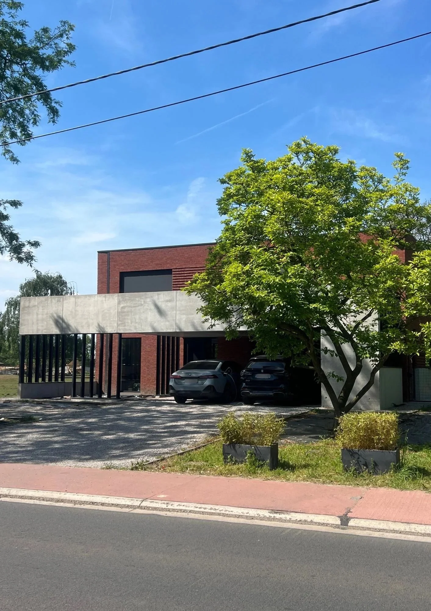 Modern house with brick and concrete facade, large garage with several parked cars, surrounded by trees and landscaping, under a clear blue sky.