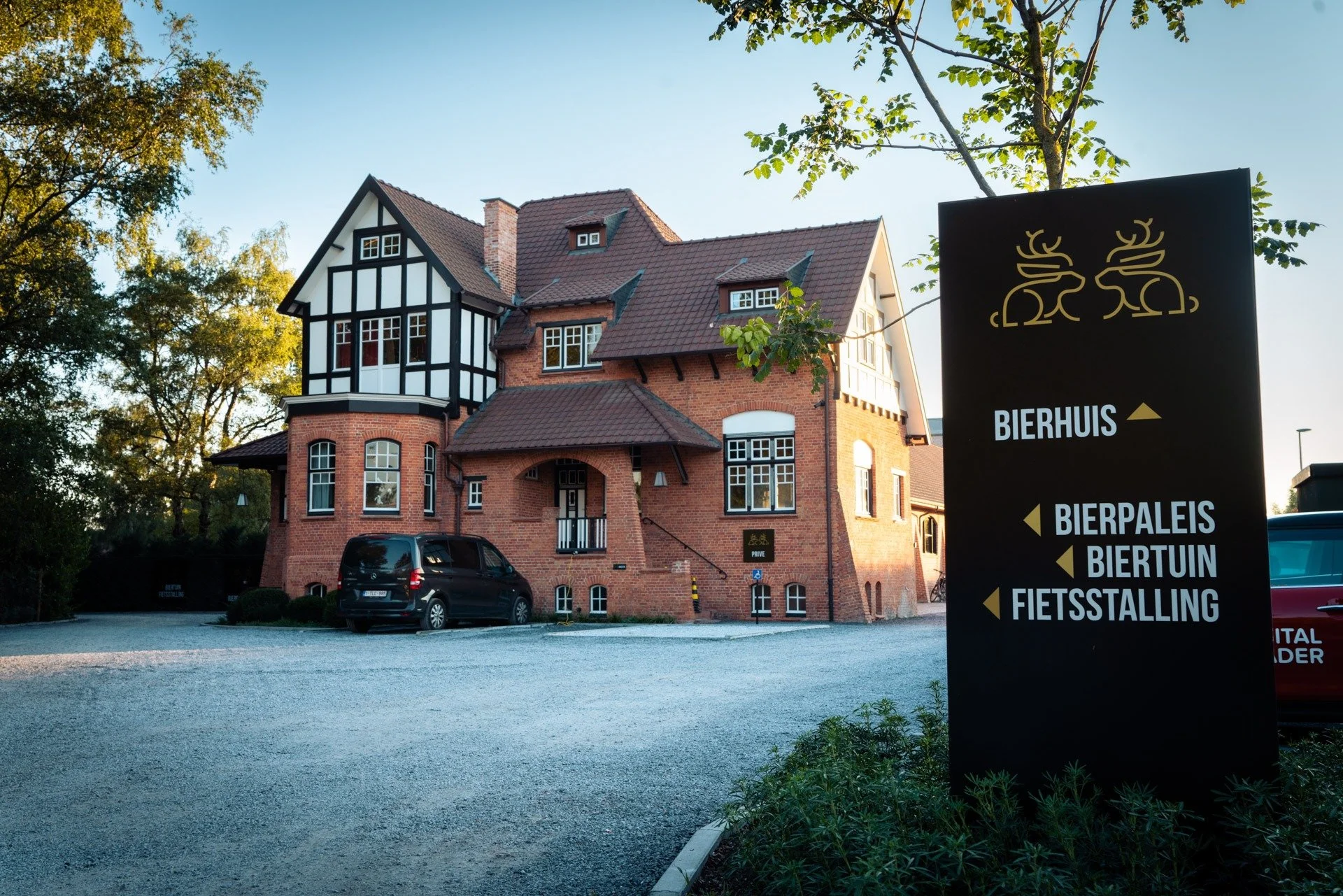 A large brick and half-timbered building with multiple windows and a red tile roof, parked cars in front, and a black sign with yellow text and symbols indicating directions to a beer house, beer garden, and other amenities, set outdoors on a clear day.
