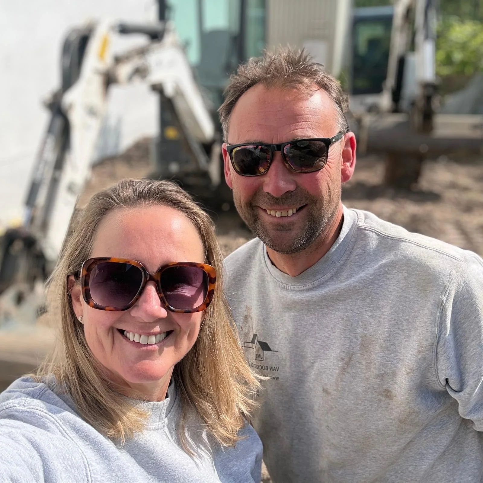 A woman and a man smiling and taking a selfie at a construction site with heavy machinery in the background.