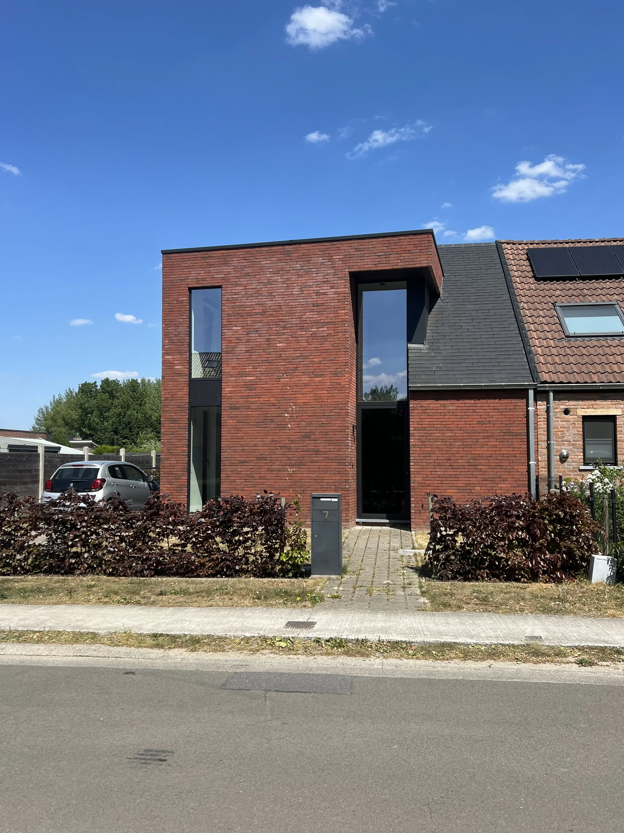 Modern red brick house with large windows, black roofing tiles, and solar panels, situated on a street with a car parked nearby, under a blue sky with some clouds and a tree in the background.