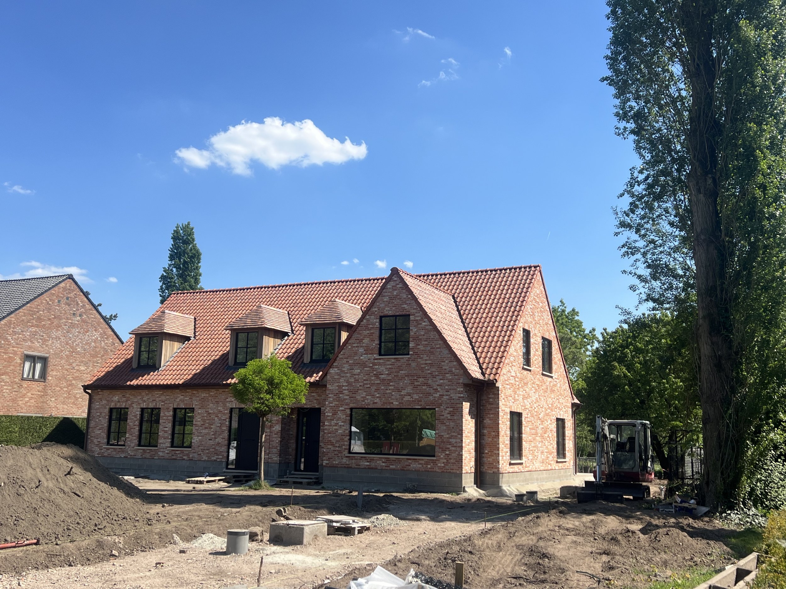 A partially constructed brick house with a red tiled roof, several large windows, and a small tree in front. Construction equipment and dirt are visible in the foreground, with a clear blue sky and some clouds overhead.