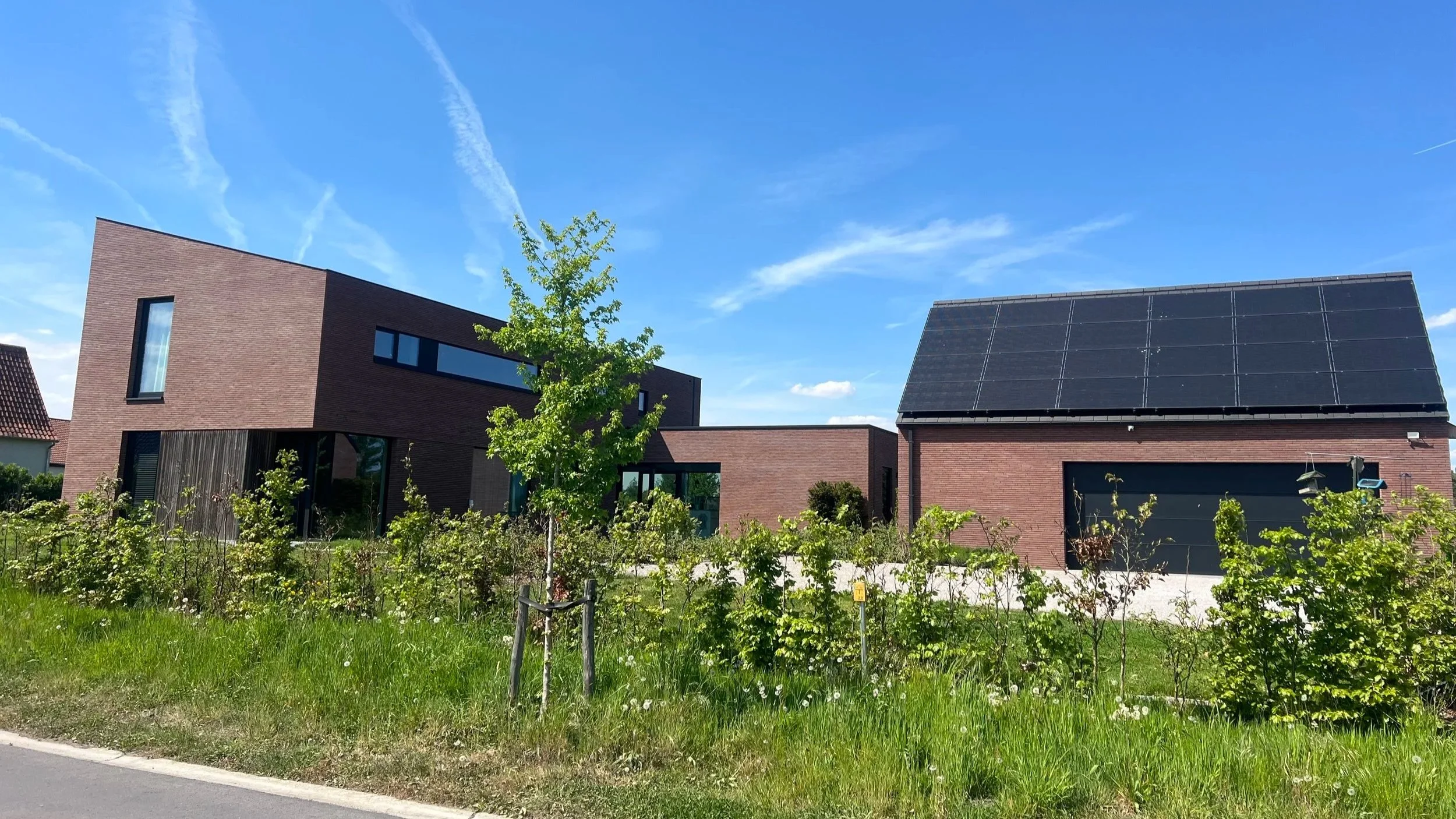 Modern brick house with solar panels on the roof, surrounded by greenery and a blue sky with clouds and contrails.