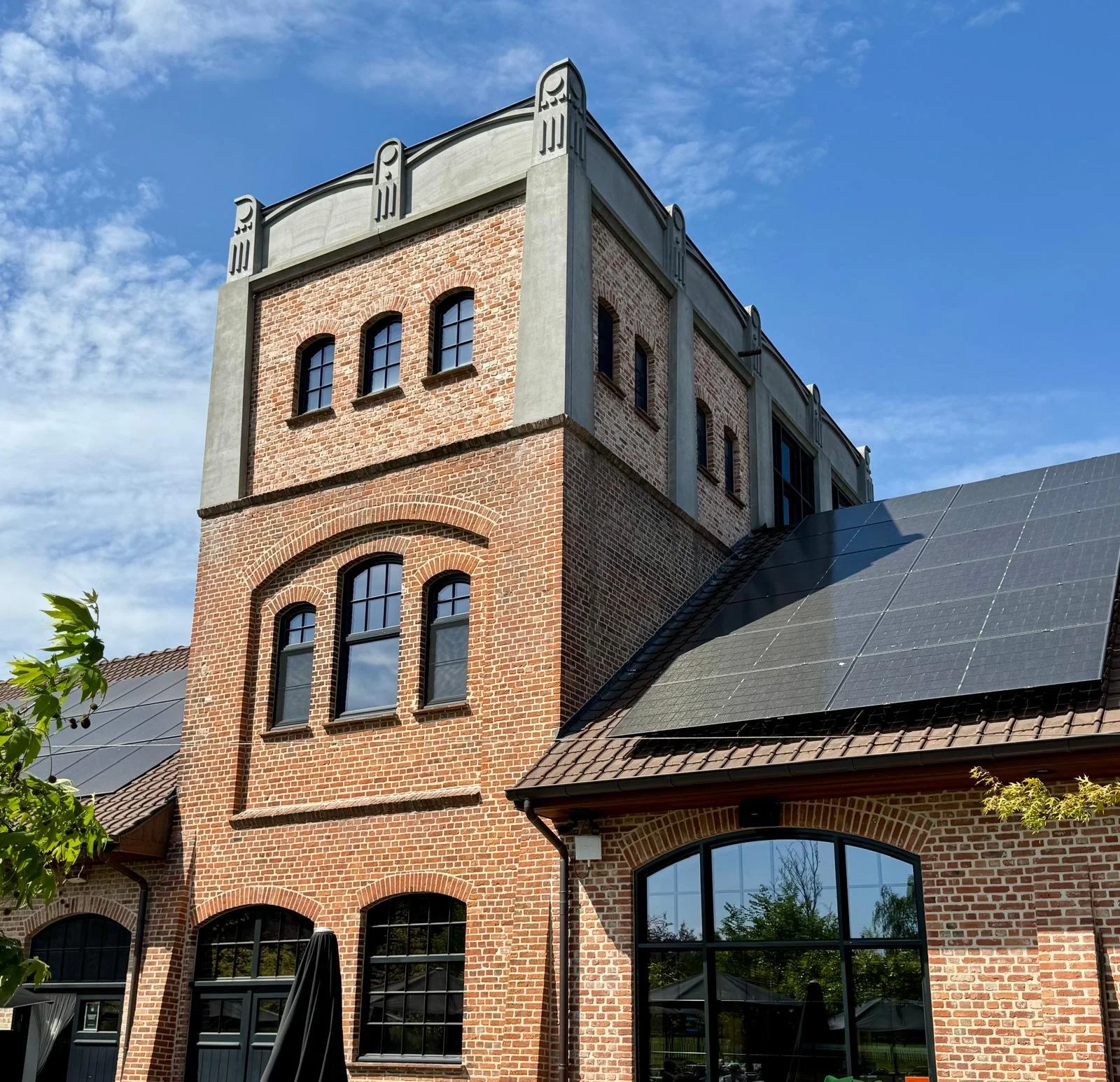 A multi-story brick building with large arched windows, a sloped roof with solar panels, and decorative concrete architectural elements around the top of the building under a blue sky with some clouds.