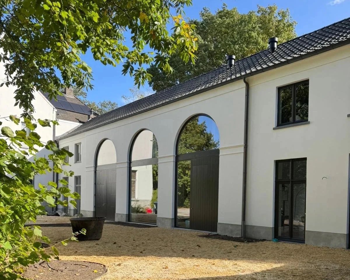 Modern white building with large arched windows and black doors, surrounded by trees and a gravel yard under a clear blue sky.