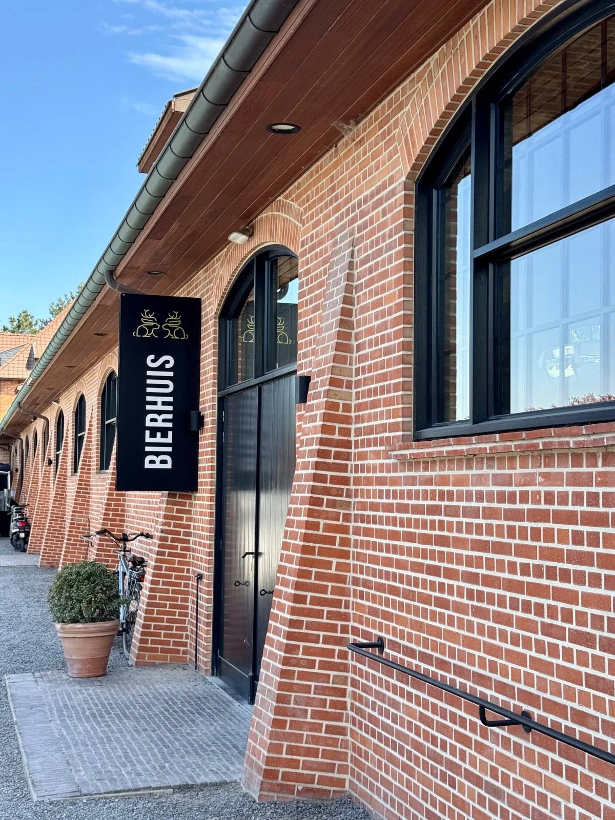 Exterior view of a brick building with a sign that reads 'Bierhuis' and large black-framed windows. There are bicycles and a potted plant near the entrance, with a cobblestone pathway in front.
