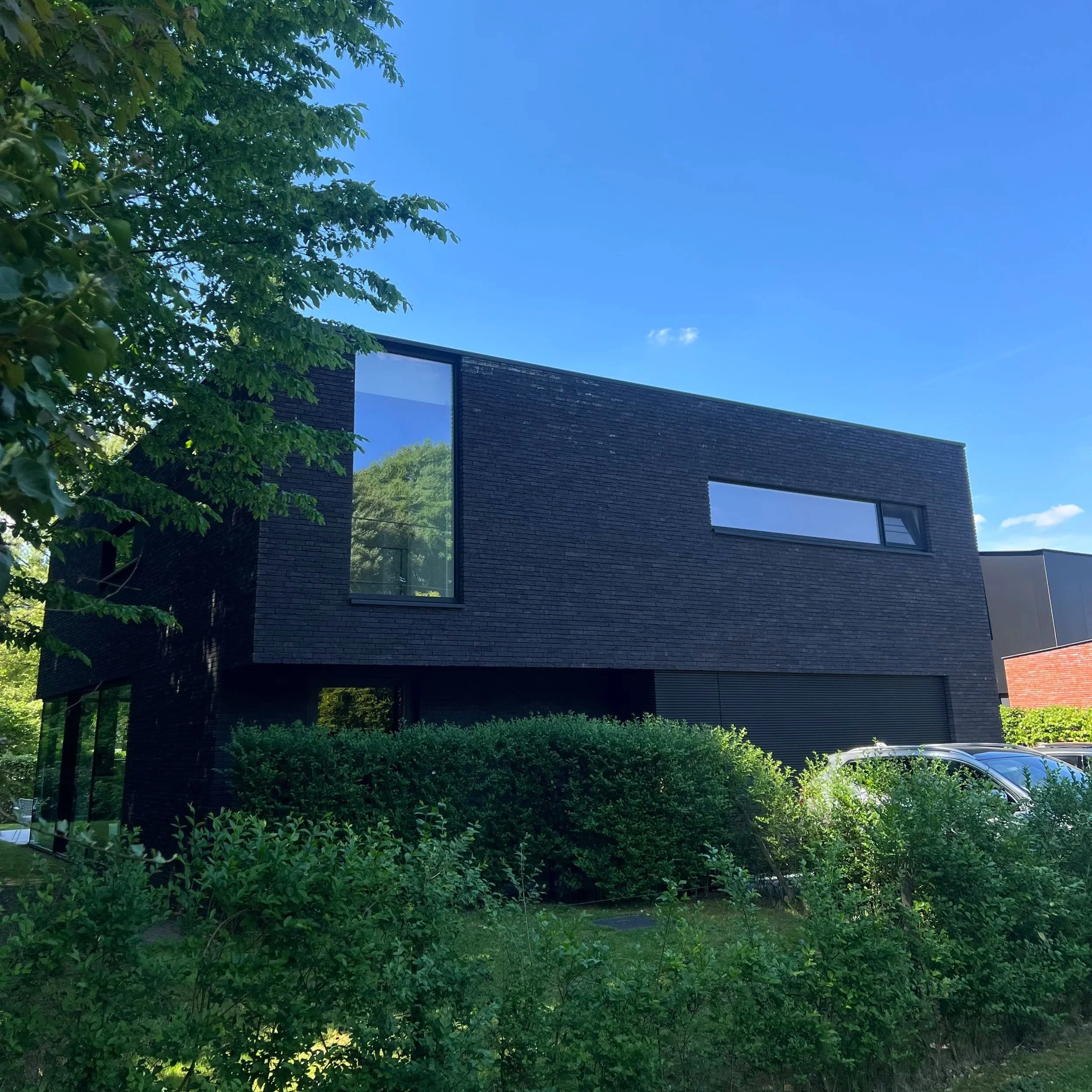 Modern building with dark brick exterior and large windows, surrounded by lush greenery and trees under a clear blue sky.